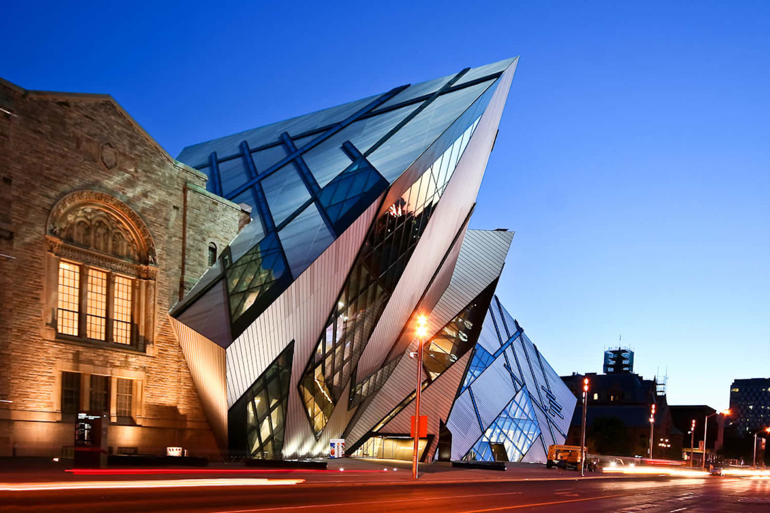 Beautiful Architecture 桌布 壁紙 — 一座引人注目的現代玻璃和鋼結構建築，帶有銳利的幾何角度，矗立在歷史悠久的石結構旁邊，背景是深藍色的暮色天空，點綴著城市燈光的條紋。