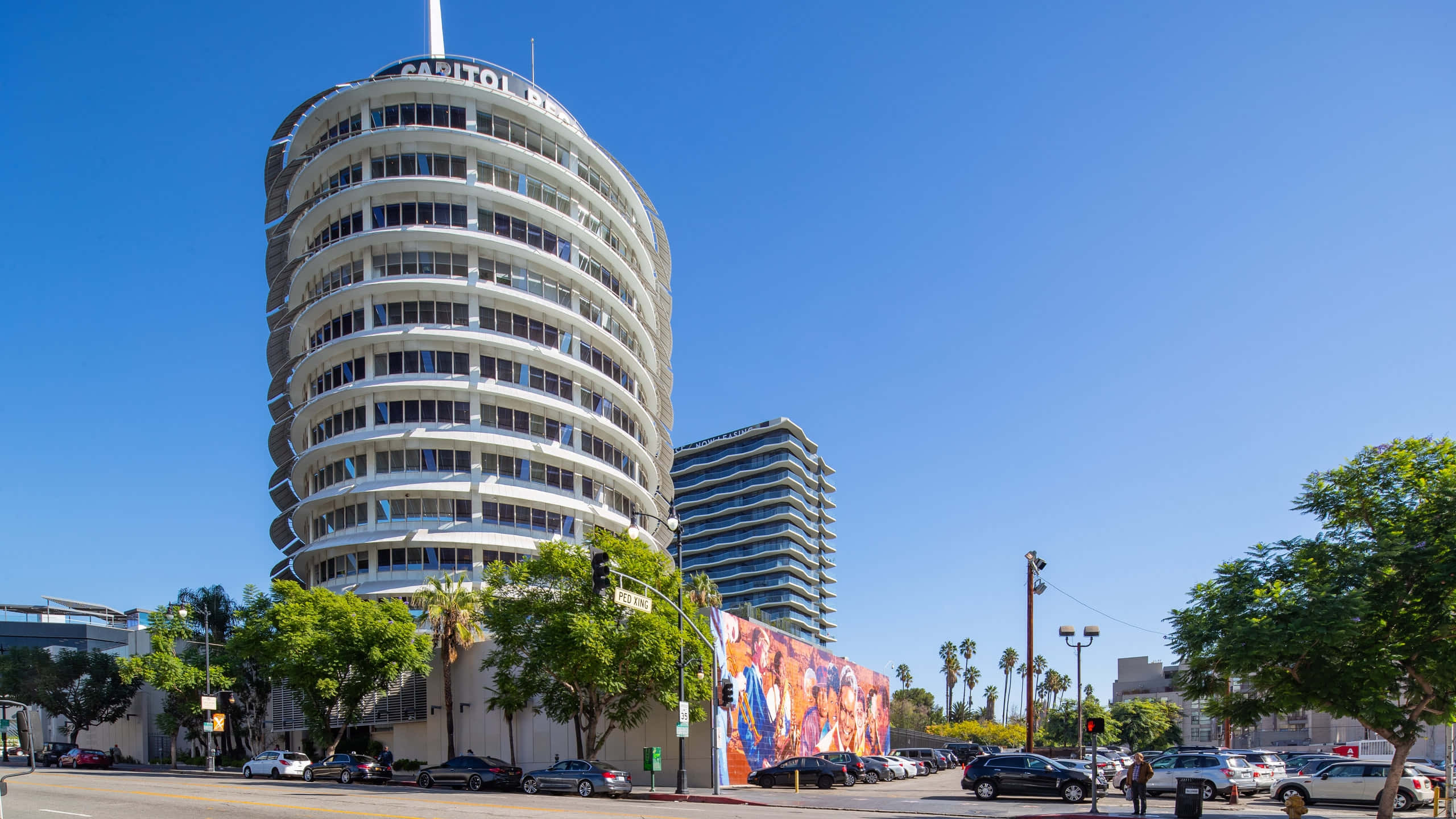 Sunny Day 桌布 壁紙 — 一座引人注目的白色弧形 Capitol Records 大樓聳立在陽光明媚、種滿棕櫚樹和停滿汽車的街道上。