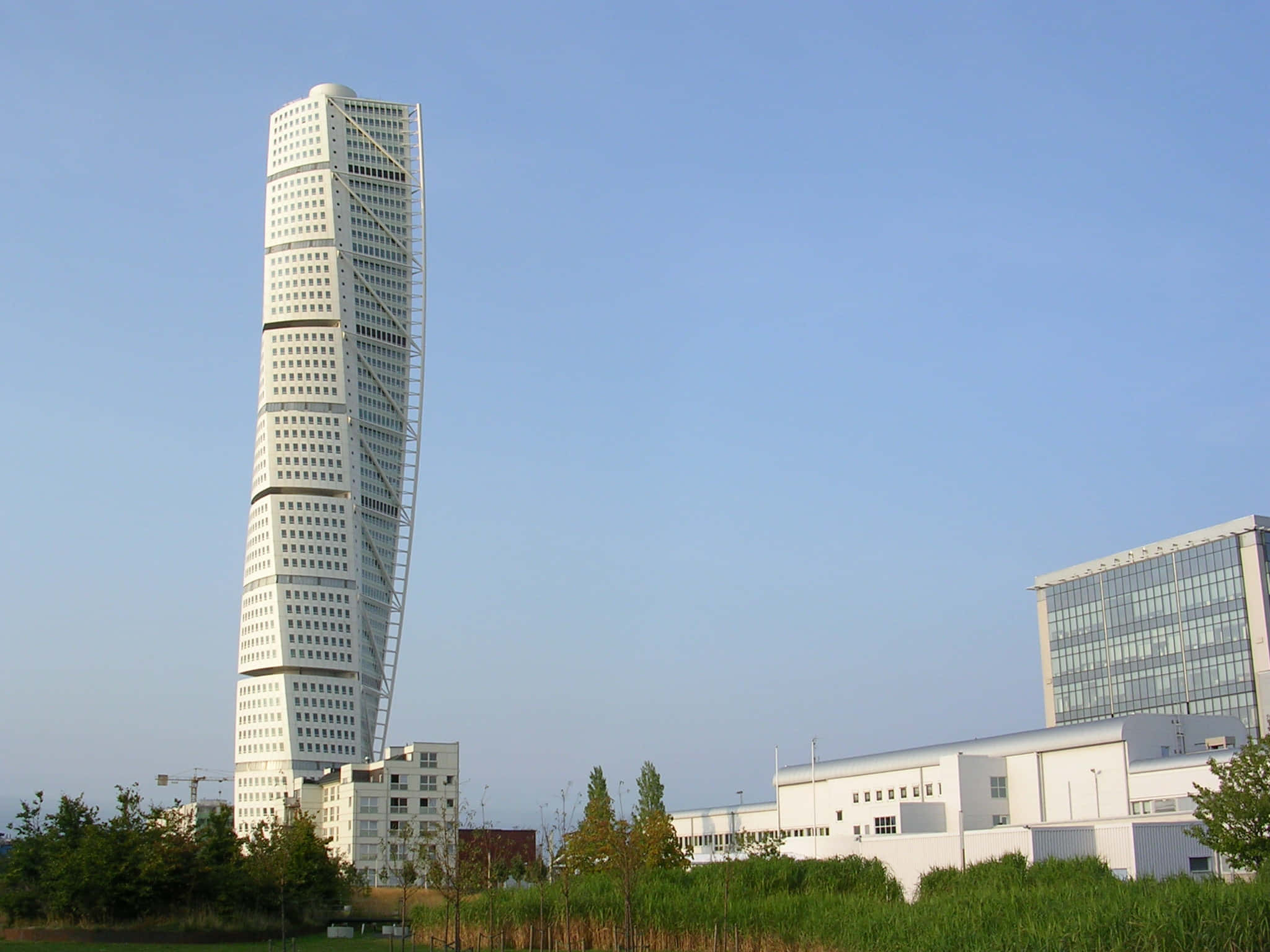 Building 桌布 壁紙 — 一座引人注目的白色扭曲摩天大樓在清澈的藍天下聳立，底部有現代建築和綠化。