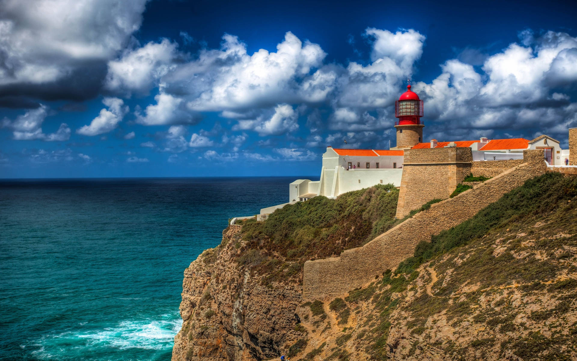 Portugal 桌布 壁紙 — 一座引人注目的紅白燈塔矗立在崎嶇的懸崖上，俯瞰著碧綠的海浪，天空蔚藍，雲朵蓬鬆。