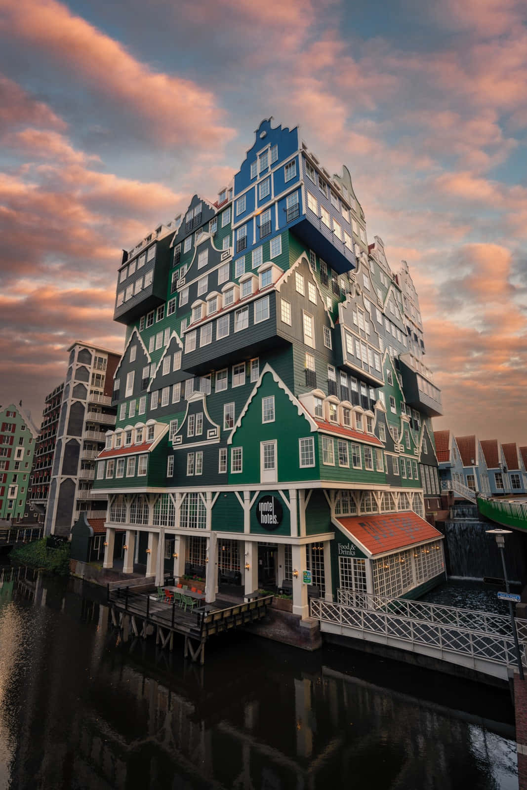 Building 桌布 壁紙 — 一座引人注目的綠色和藍色現代酒店，像荷蘭山牆房屋一樣堆疊，在戲劇性的日落天空下倒映在平靜的運河水中。