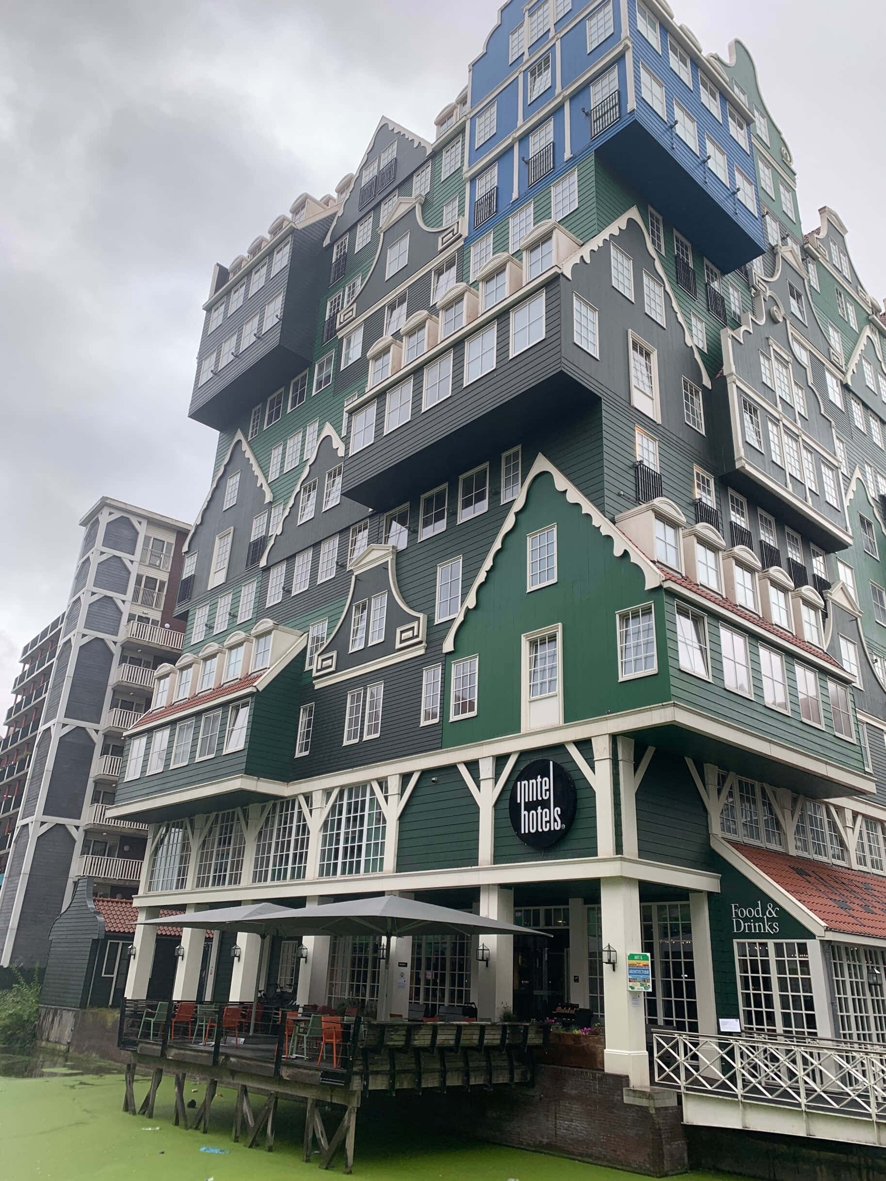建築 桌布 壁紙 — 一座引人注目的綠色和藍色階梯式山牆酒店建築，帶有白色飾邊，矗立在綠色運河之上，天空陰沉。