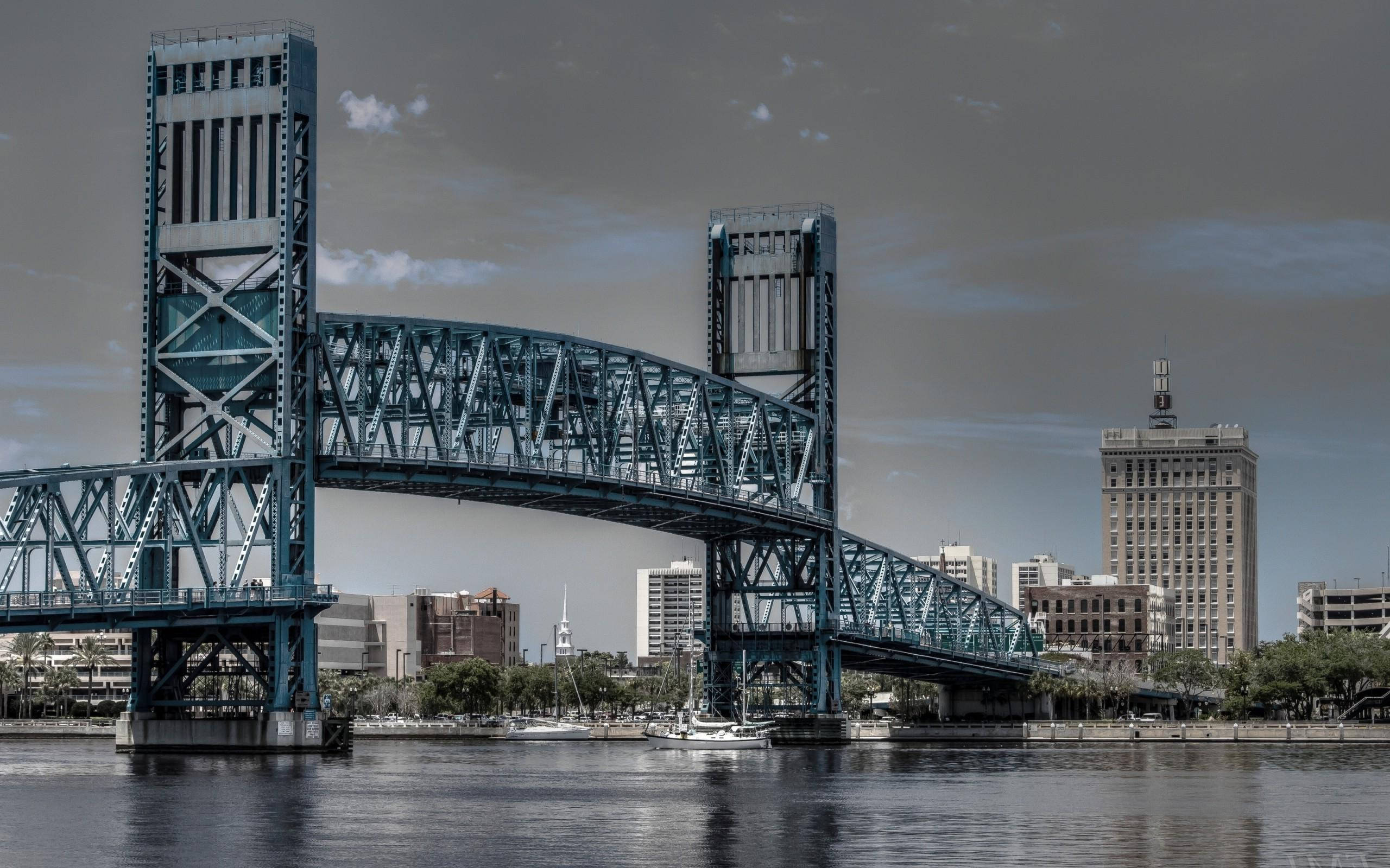 Jacksonville 桌布 壁紙 — 一座引人注目的藍色鋼桁架橋橫跨平靜的河流，背景中城市建築在陰沉的灰色天空下聳立。
