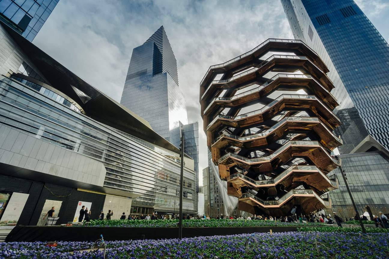New York 桌布 壁紙 — 一座引人注目的螺旋木結構在玻璃摩天大樓中升起，其底部在陰天天空下盛開著紫色花朵。