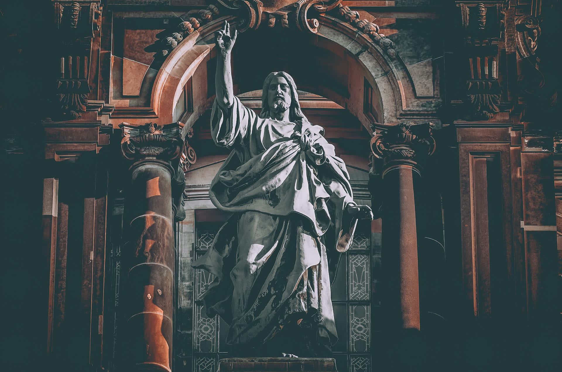 Catholic Desktop 桌布 壁紙 — 一座戲劇性的耶穌基督雕像，舉起手，站在光線昏暗的大教堂內一個華麗的拱形門口下。