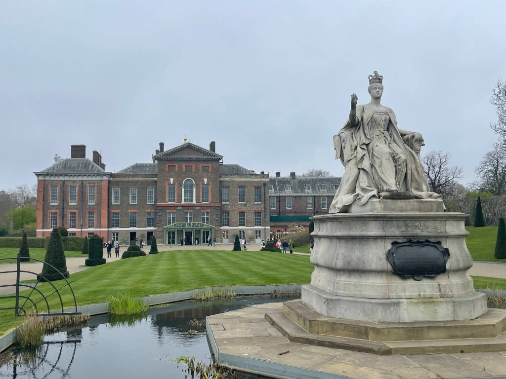 England 桌布 壁紙 — 一座戴著王冠的女王大型石像矗立在一棟莊嚴的磚造豪宅前，擁有修剪整齊的花園和一個在陰天天空下的倒影池。