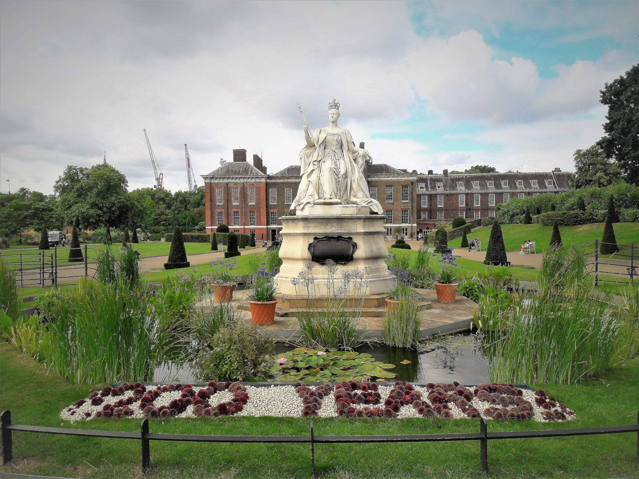 England 桌布 壁紙 — 一座戴著王冠的女王白色大雕像矗立在一個有睡蓮池塘和「300年」花卉展示的正式花園中。