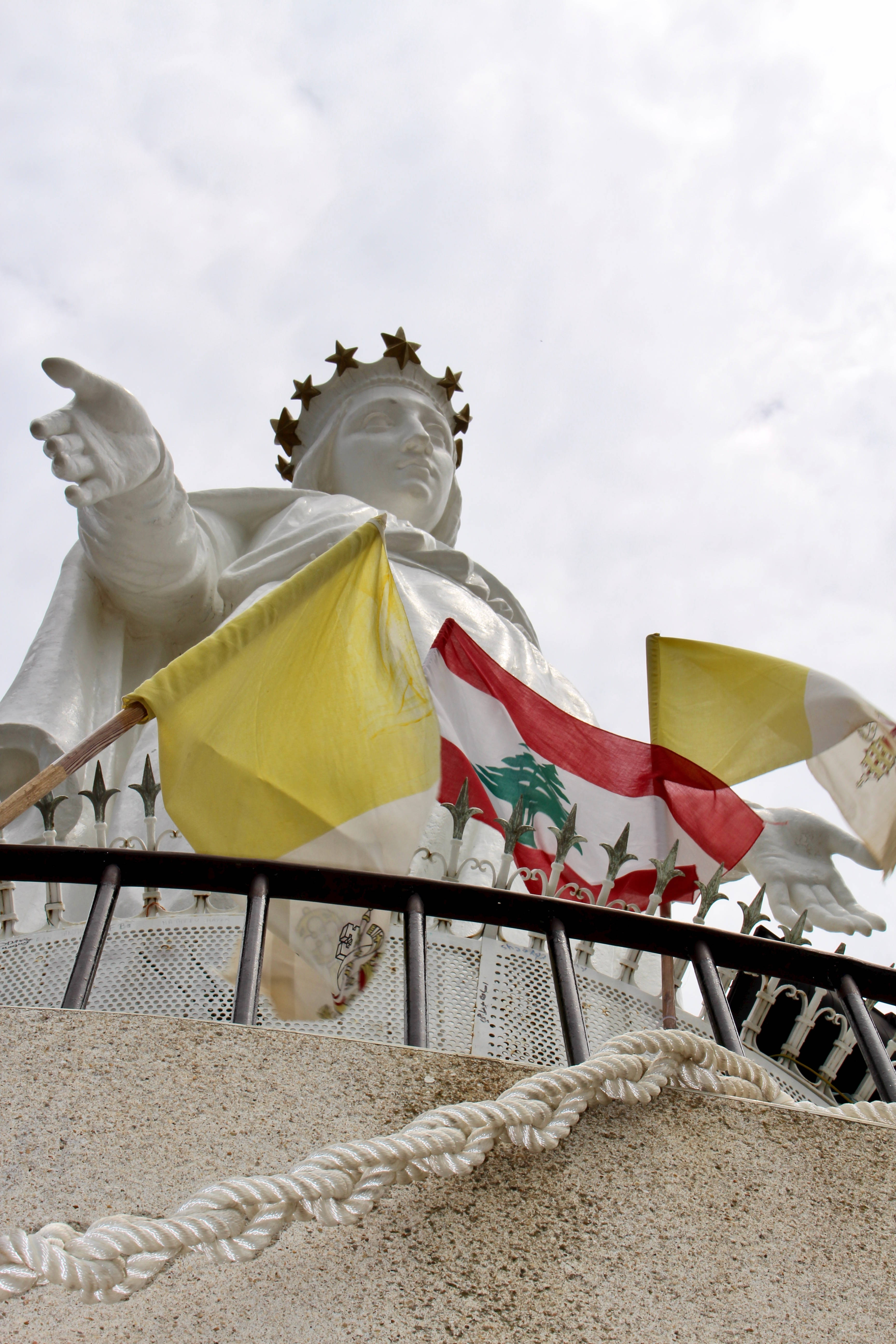 Lebanon 桌布 壁紙 — 一座戴著皇冠的聖母瑪利亞白色雕像高聳入雲，手指向前方，黎巴嫩和梵蒂岡旗幟在前方飄揚。