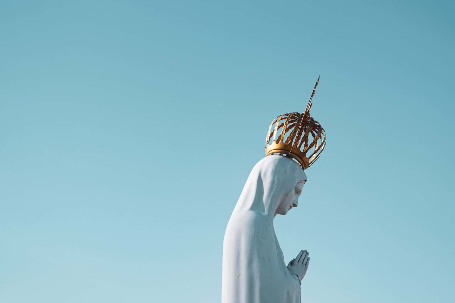 Mother Mary 桌布 壁紙 — 一座戴著金色皇冠的聖母瑪利亞白色雕像，在清澈的藍天下祈禱。