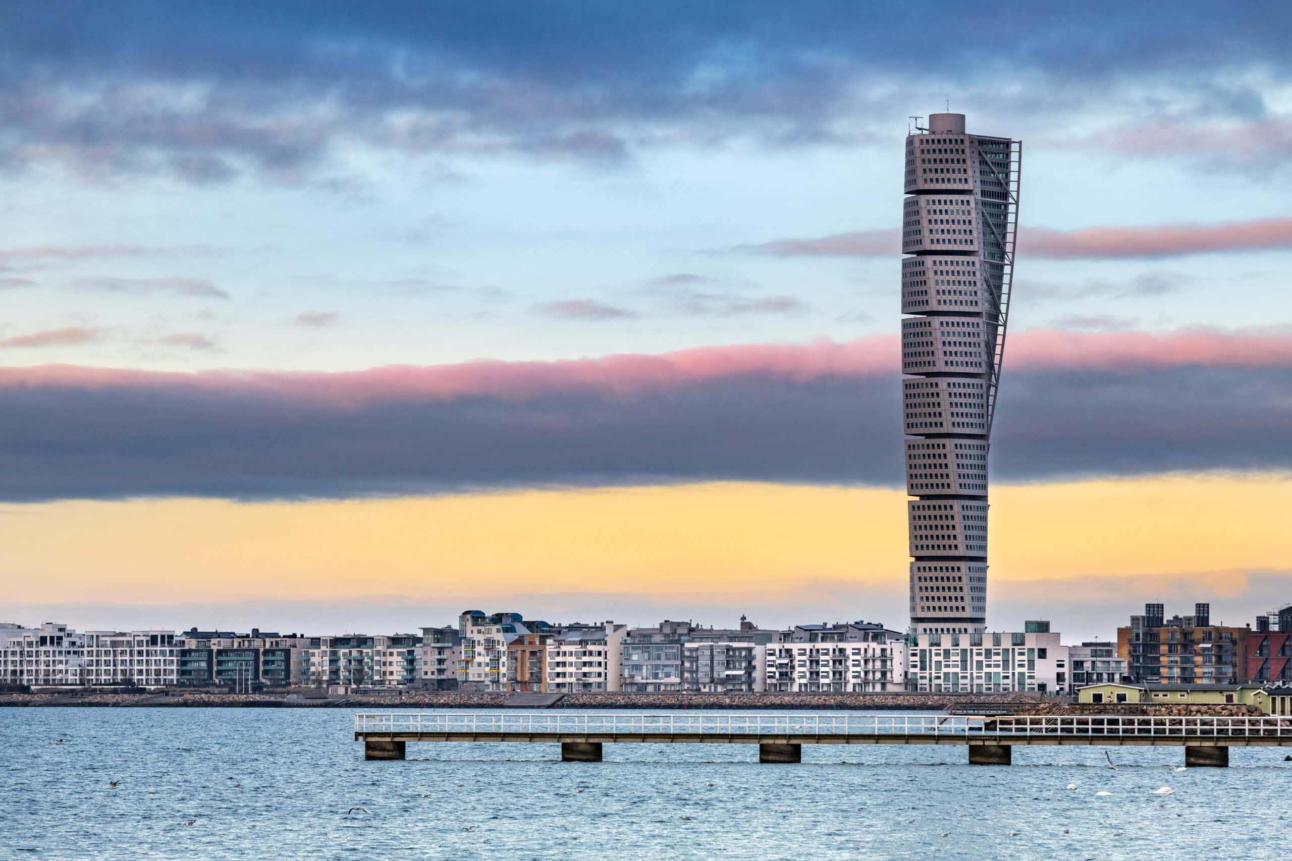Skyscraper 桌布 壁紙 — 一座扭曲的現代摩天大樓從海濱城市景觀中升起，夕陽下粉紅色和藍色的雲朵映照在平靜的水面上。