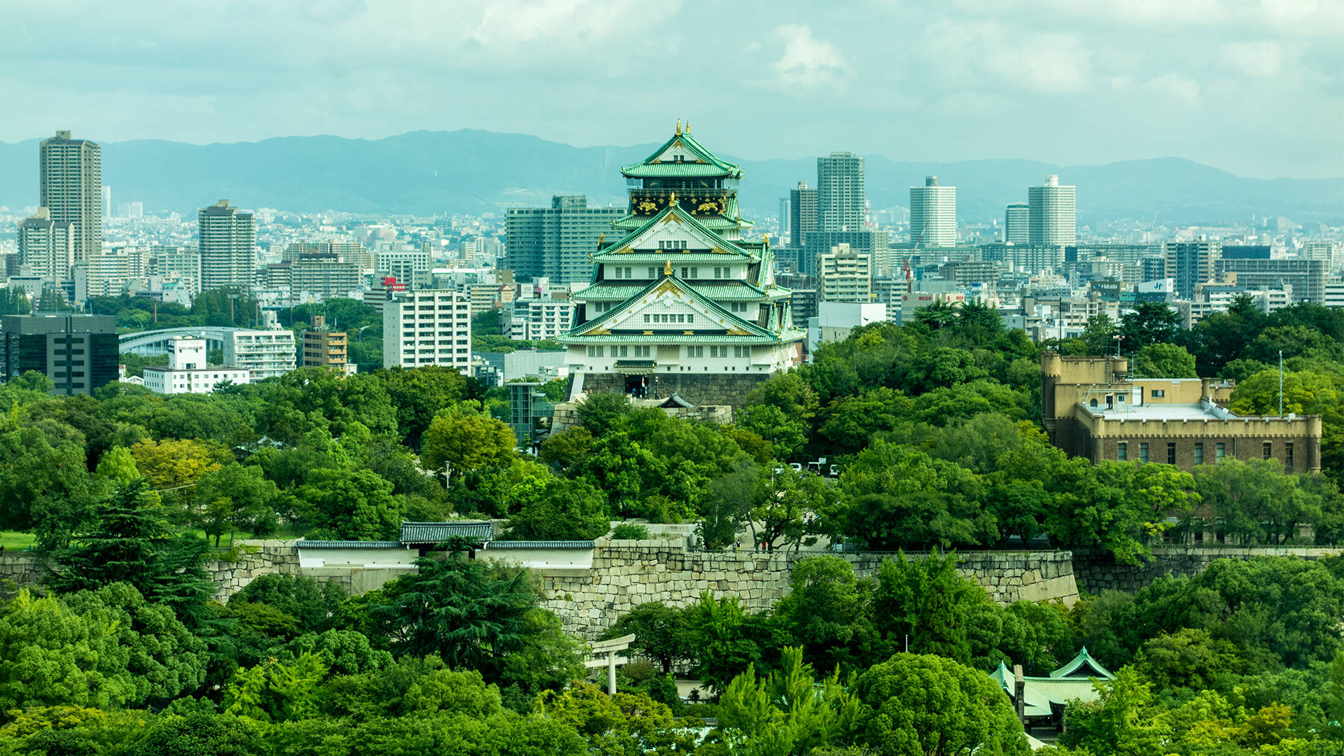 Osaka Castle 桌布 壁紙 — 一座擁有綠色屋頂的宏偉傳統日本城堡坐落在鬱鬱蔥蔥的山丘上，俯瞰著多雲天空下的現代城市景觀。