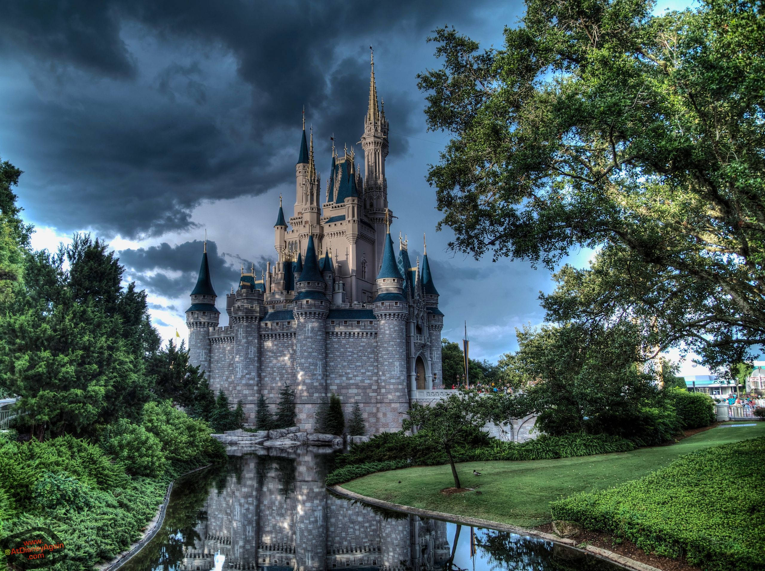 Disneyland Castle 桌布 壁紙 — 一座擁有藍色尖頂的雄偉城堡矗立在暴風雨的天空下，倒映在平靜的護城河中，四周環繞著茂密的綠樹。