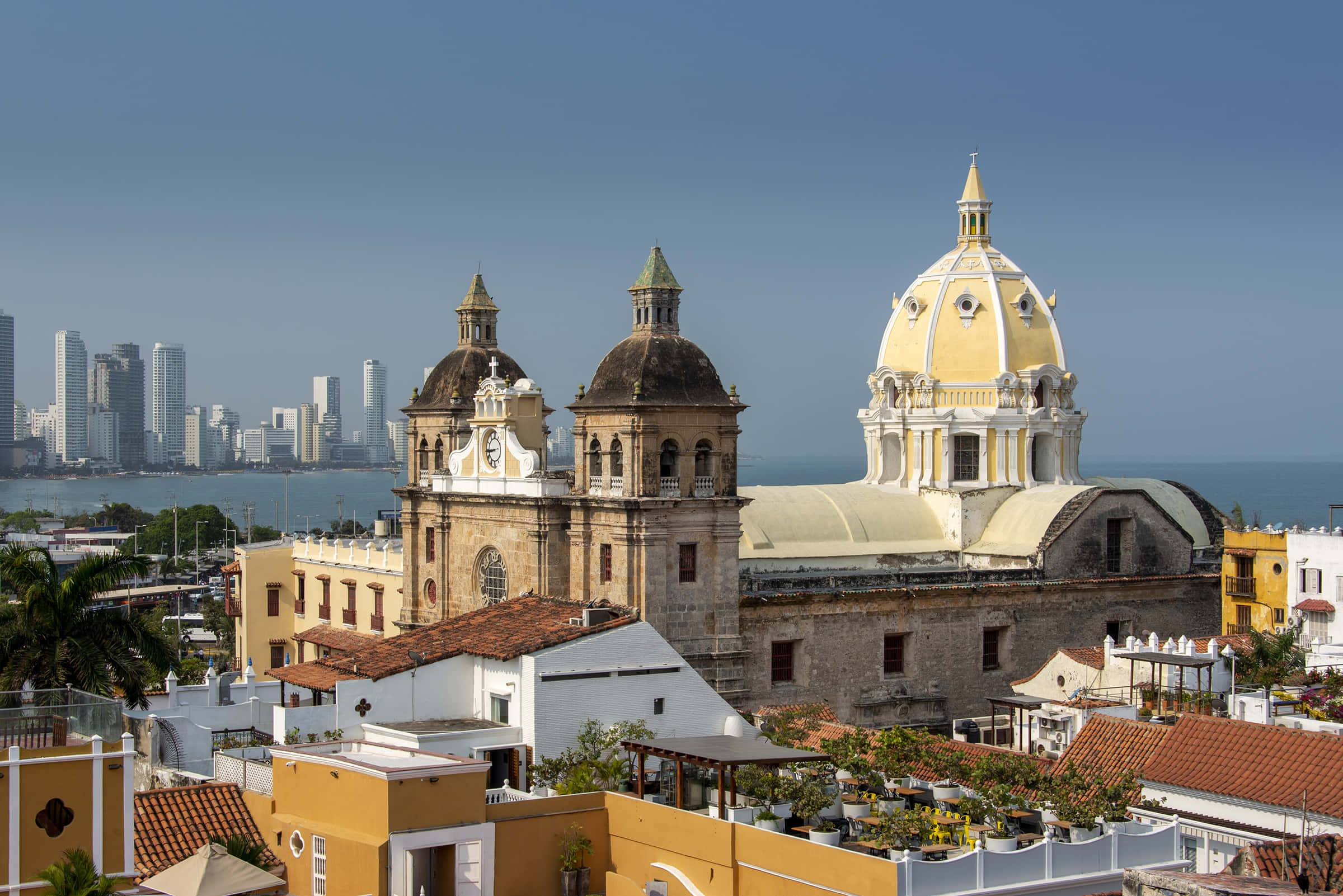 Colombia 桌布 壁紙 — 一座擁有金色圓頂的歷史大教堂矗立在色彩繽紛的殖民時期建築中，俯瞰著現代城市天際線和海洋。