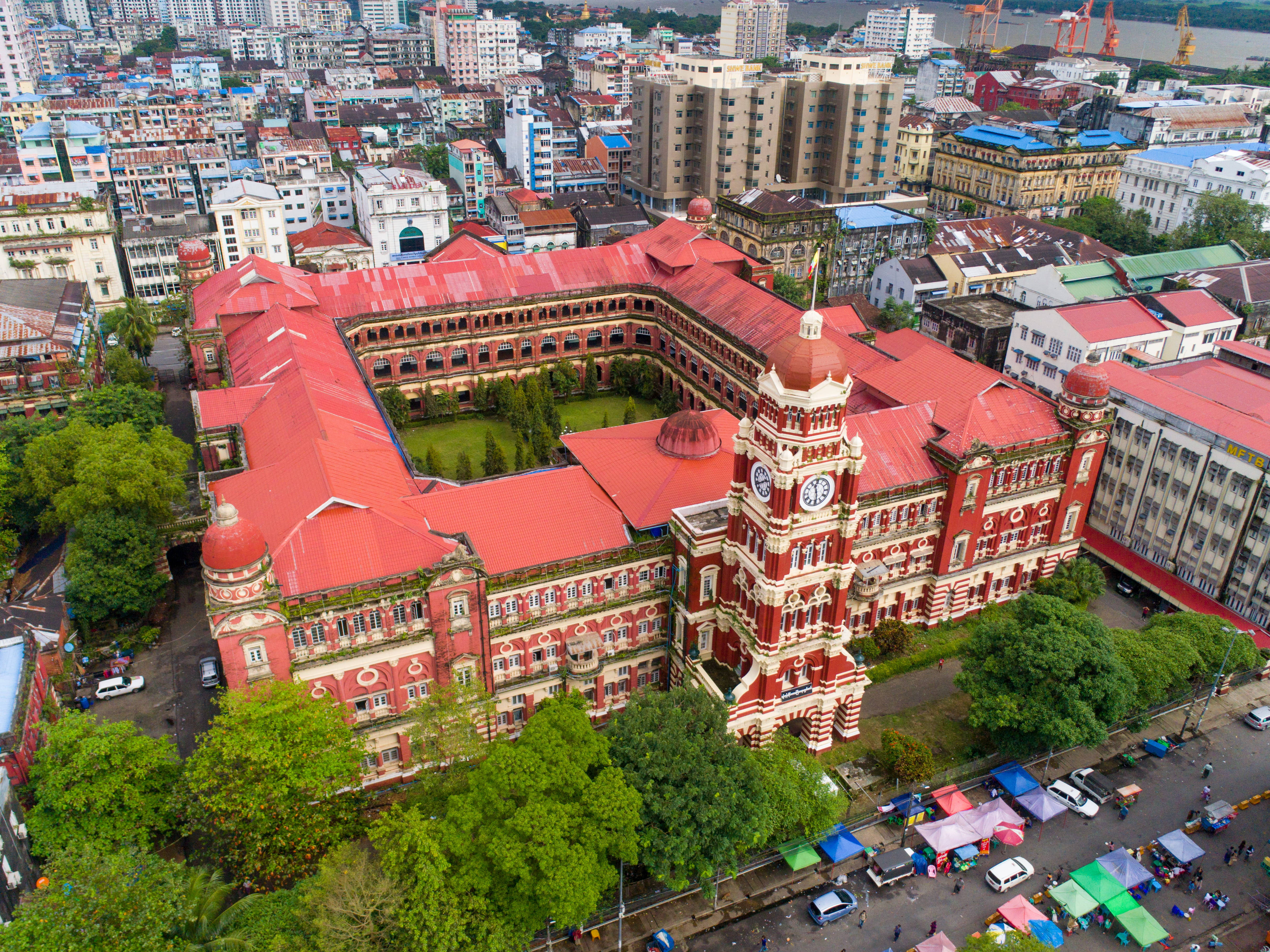 Yangon 桌布 壁紙 — 一座擁有鐘樓的宏偉紅頂殖民風格建築的鳥瞰圖，周圍環繞著茂密的綠樹和繁華的城市景觀。