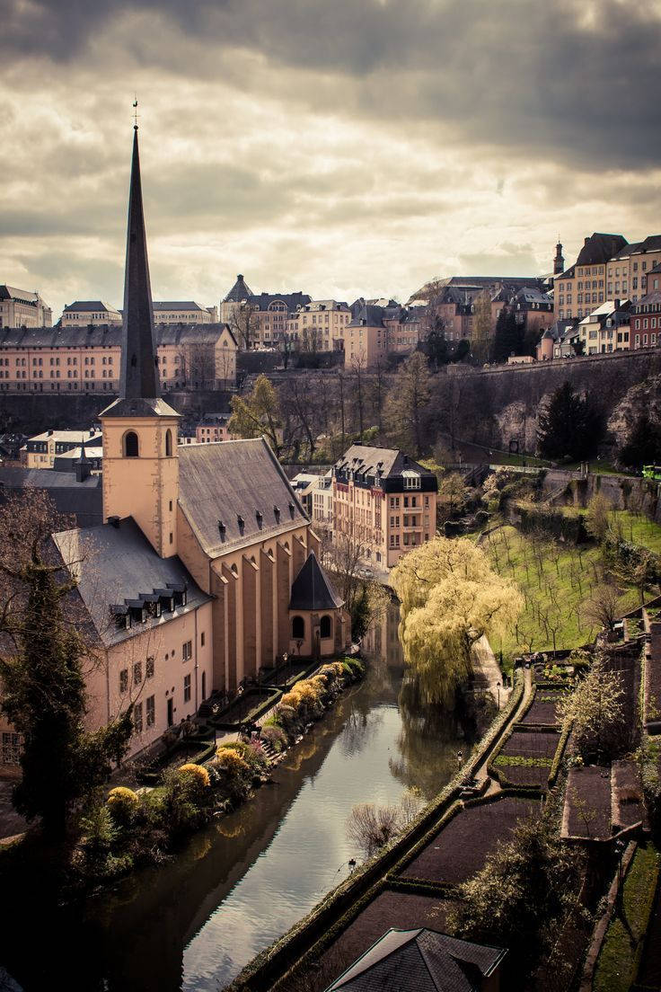 Luxembourg 桌布 壁紙 — 一座擁有高尖頂的歷史教堂矗立在平靜的運河旁，倒映著秋日的樹木和周圍的歐洲建築，天空多雲。