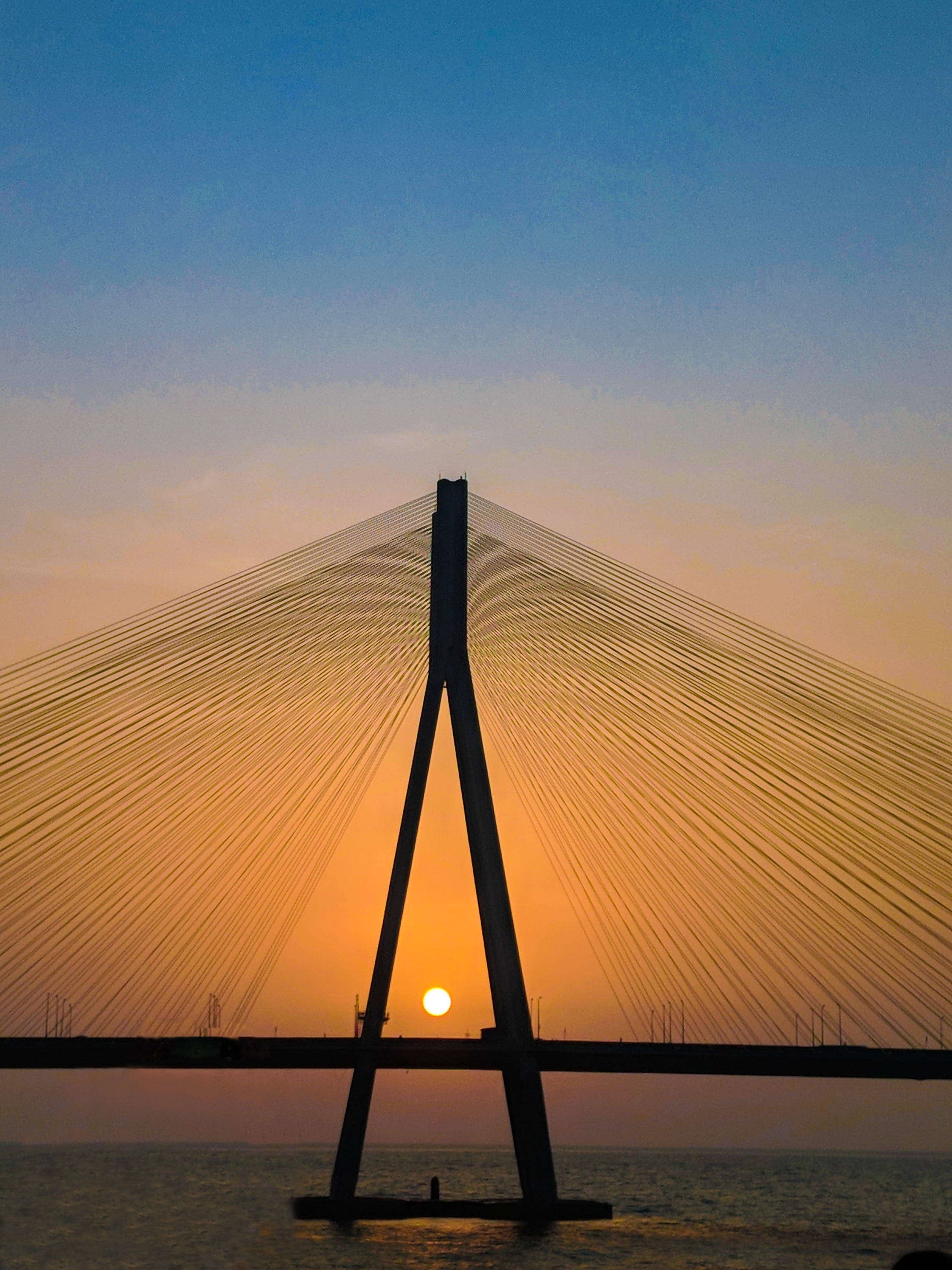Mumbai 桌布 壁紙 — 一座斜張橋在平靜的水面上，以中央橋塔背後發光的夕陽為背景，形成剪影。
