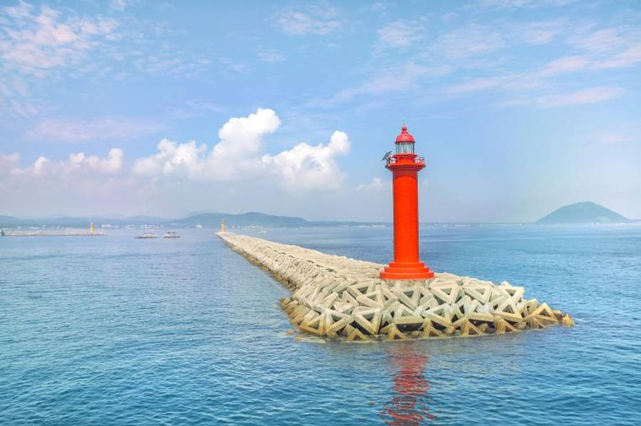 Jeju 桌布 壁紙 — 一座明亮的紅色燈塔矗立在長長的石頭防波堤上，延伸至平靜的藍色大海，天空部分多雲。