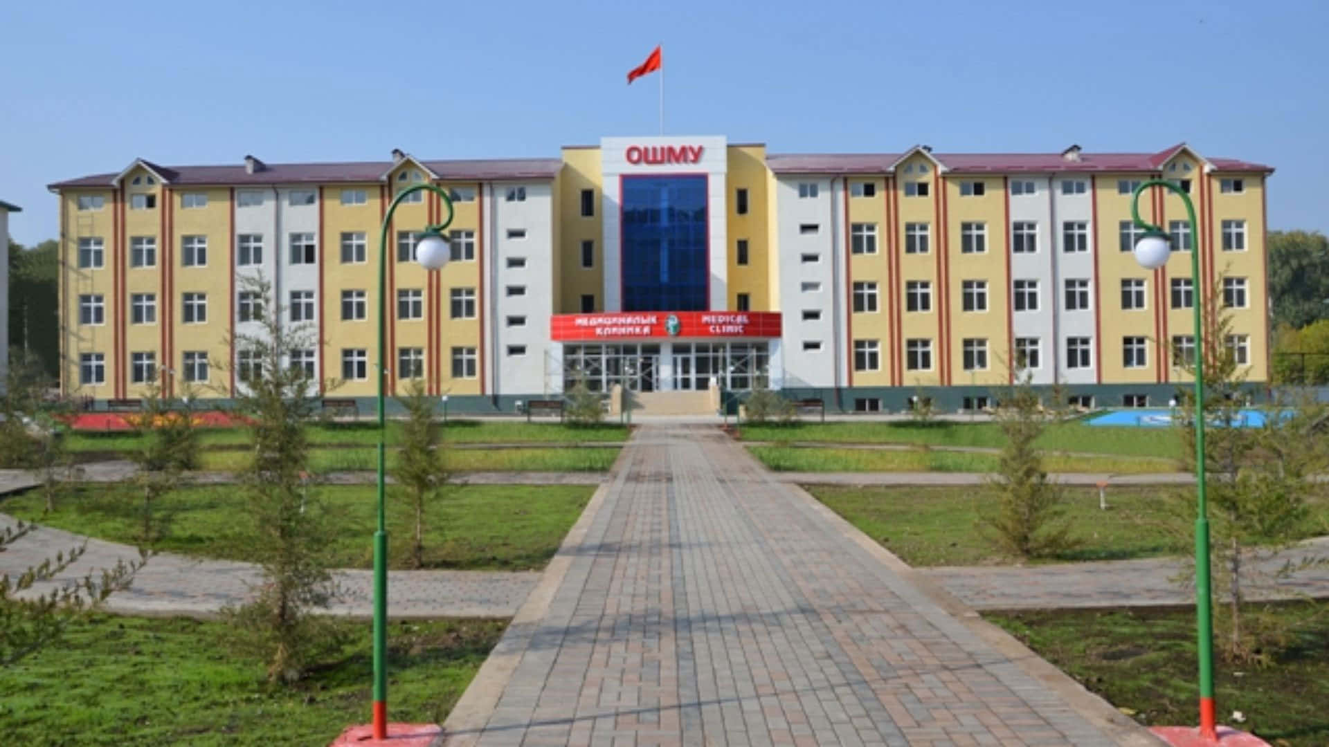 Building 桌布 壁紙 — 一座明亮的黃白相間多層建築，帶有紅色點綴，矗立在清澈的藍天下，可通過一條鋪砌的小路抵達，小路兩旁種有幼樹和綠草。