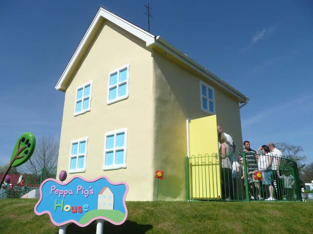 Peppa Pig Family 桌布 壁紙 — 一座明亮的黃色Peppa Pig房屋複製品，帶有藍色窗戶，矗立在晴朗的藍天下，遊客聚集在綠色籬笆附近。