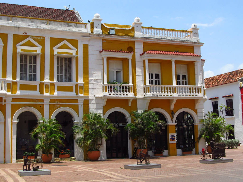 Colombia 桌布 壁紙 — 一座明亮的黃色殖民風格建築，帶有白色飾邊和紅色瓦片屋頂，矗立在晴朗的藍天下，位於陽光普照的廣場。