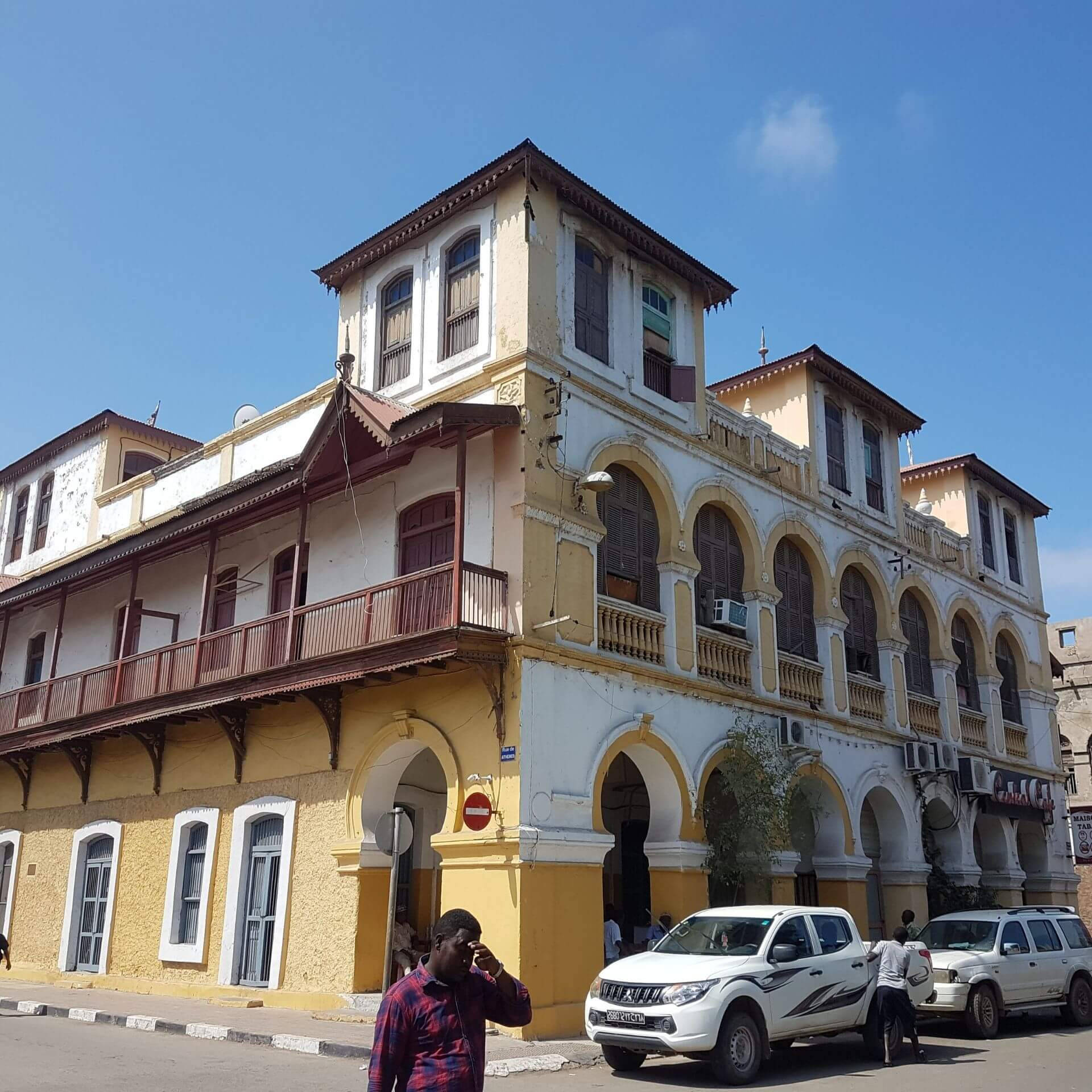 建築 桌布 壁紙 — 一座明亮的黃色殖民風格建築，帶有拱形門廊和陽台，矗立在清澈的藍天下，行人與白色SUV沿街移動。