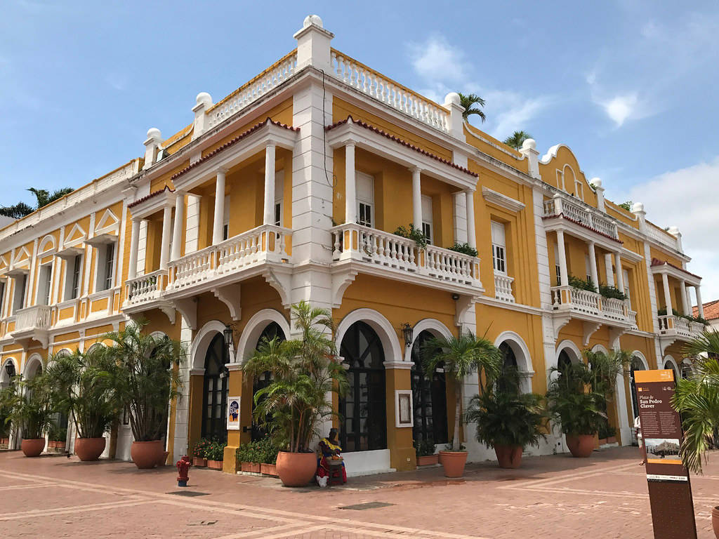 Colombia 桌布 壁紙 — 一座明亮的黃色殖民風格建築，帶有白色陽台和拱形門口，矗立在晴朗的藍天下，兩旁是磚砌廣場上的盆栽棕櫚樹。
