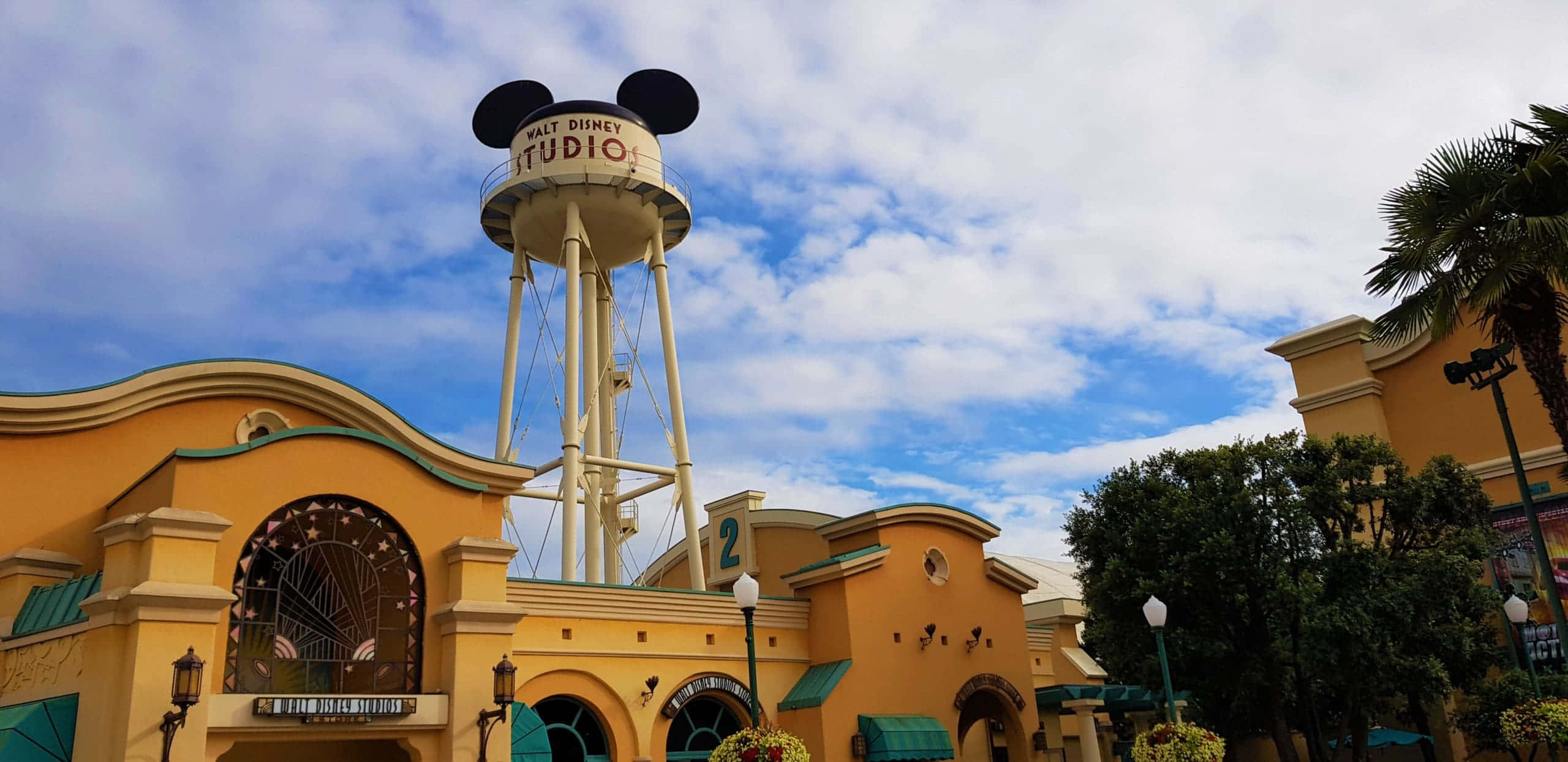 Walt Disney Studios Motion 桌布 壁紙 — 一座明亮的黃色華特迪士尼工作室水塔，帶有米奇老鼠耳朵，在主題建築上方部分多雲的藍天下高高聳立。
