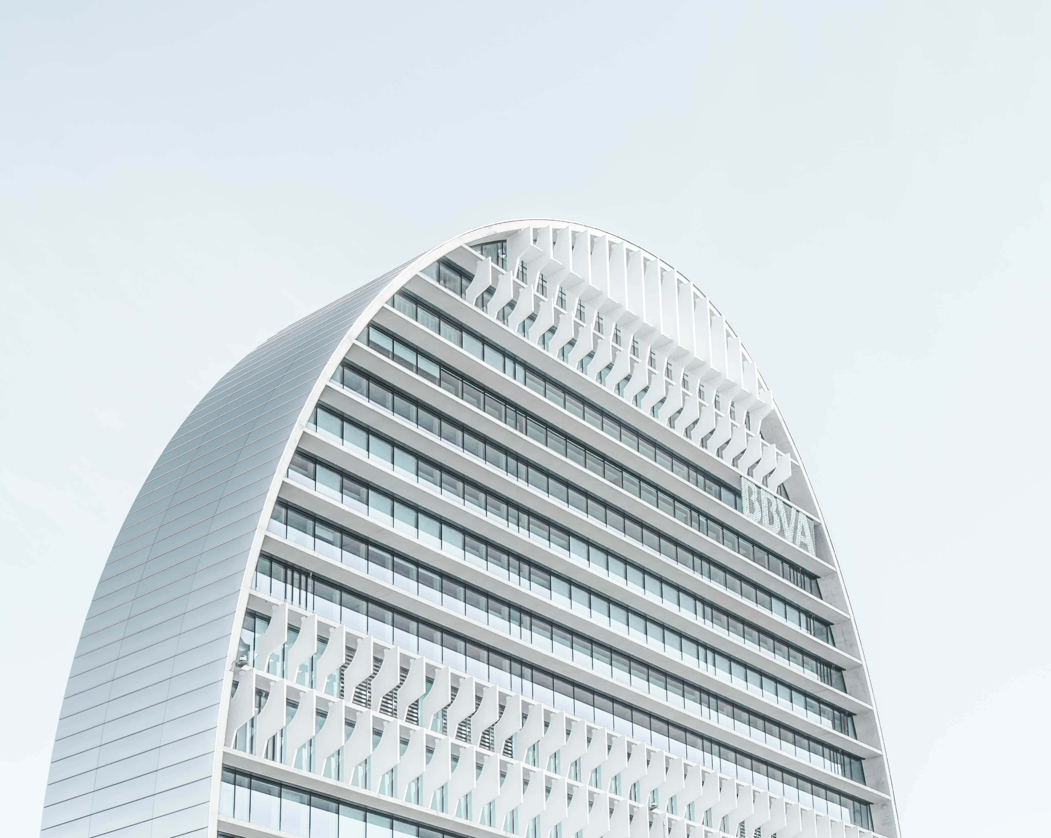 Building 桌布 壁紙 — 一座時尚現代白色彎曲摩天大樓，帶有玻璃窗和BBVA標誌，在淡色天空下聳立。