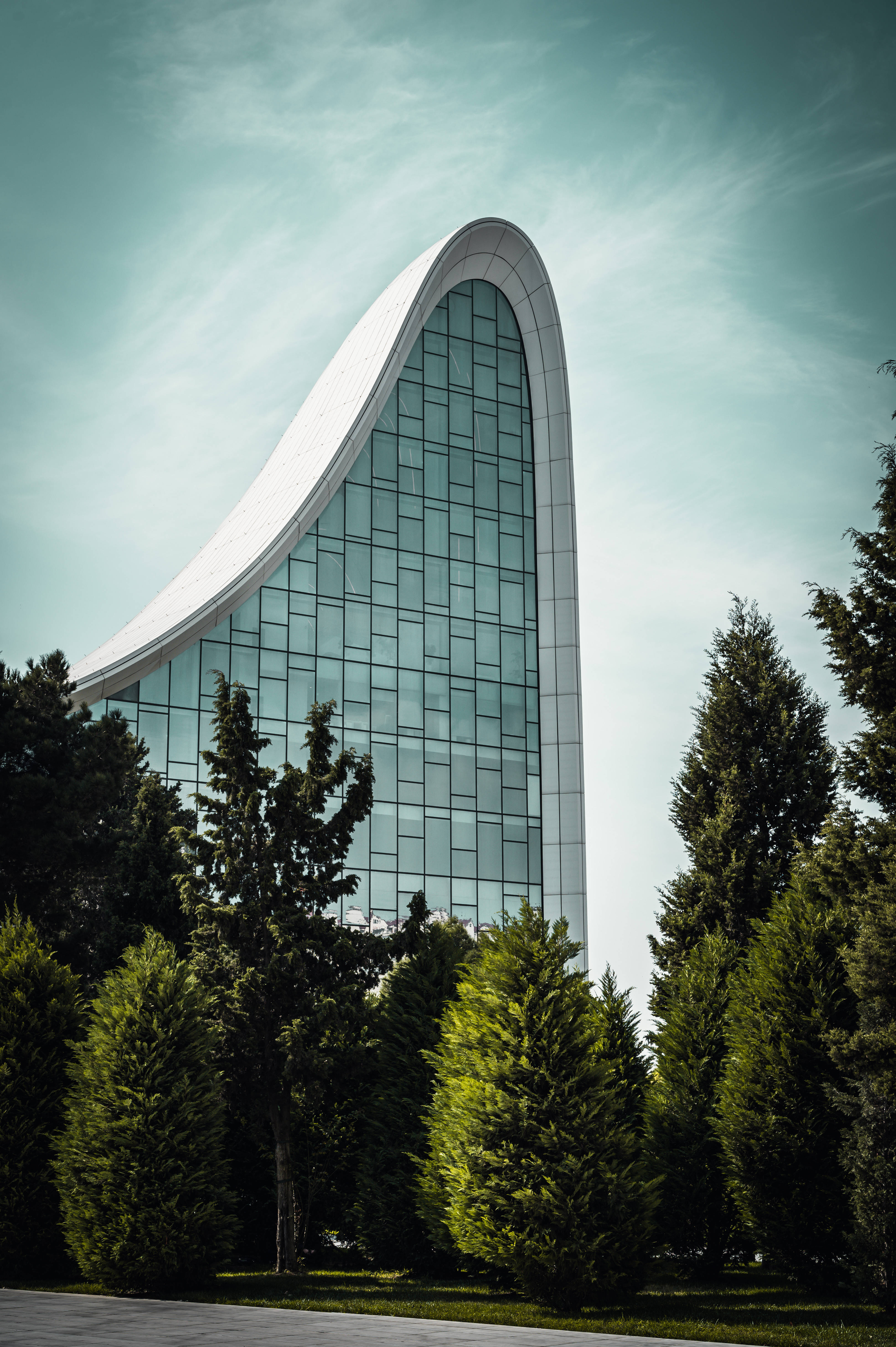 Azerbaijan 桌布 壁紙 — 一座時尚的現代建築，擁有弧形玻璃外牆，在淡藍色天空下聳立在茂密的綠樹之上。