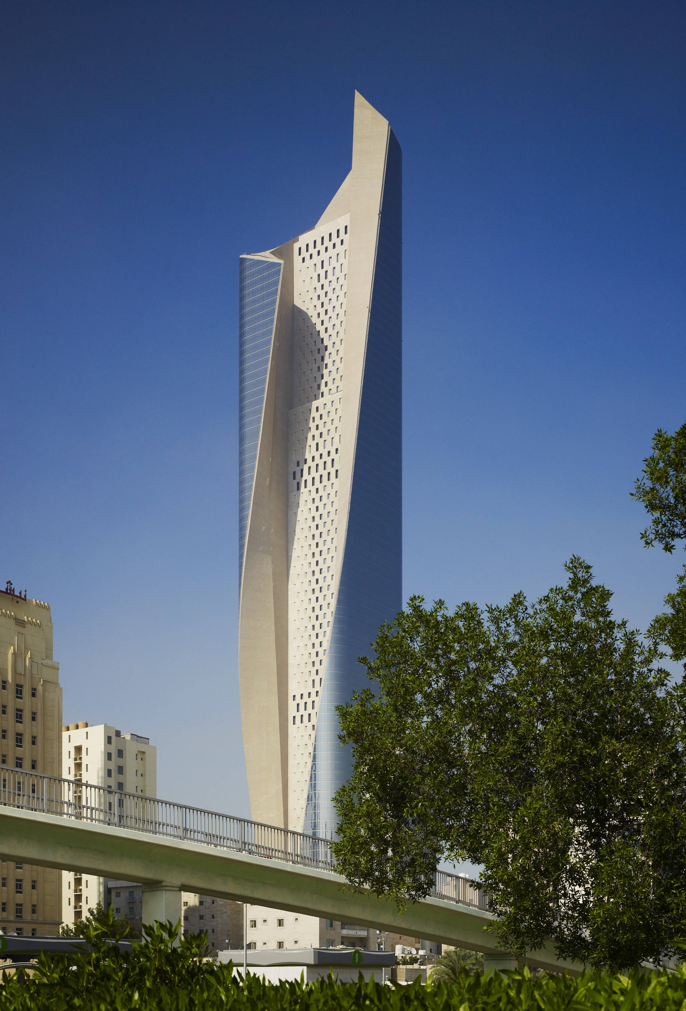 Kuwait 桌布 壁紙 — 一座時尚的現代摩天大樓，擁有扭曲的玻璃和混凝土外牆，在藍天白雲下聳立，周圍環繞著綠樹和人行天橋。