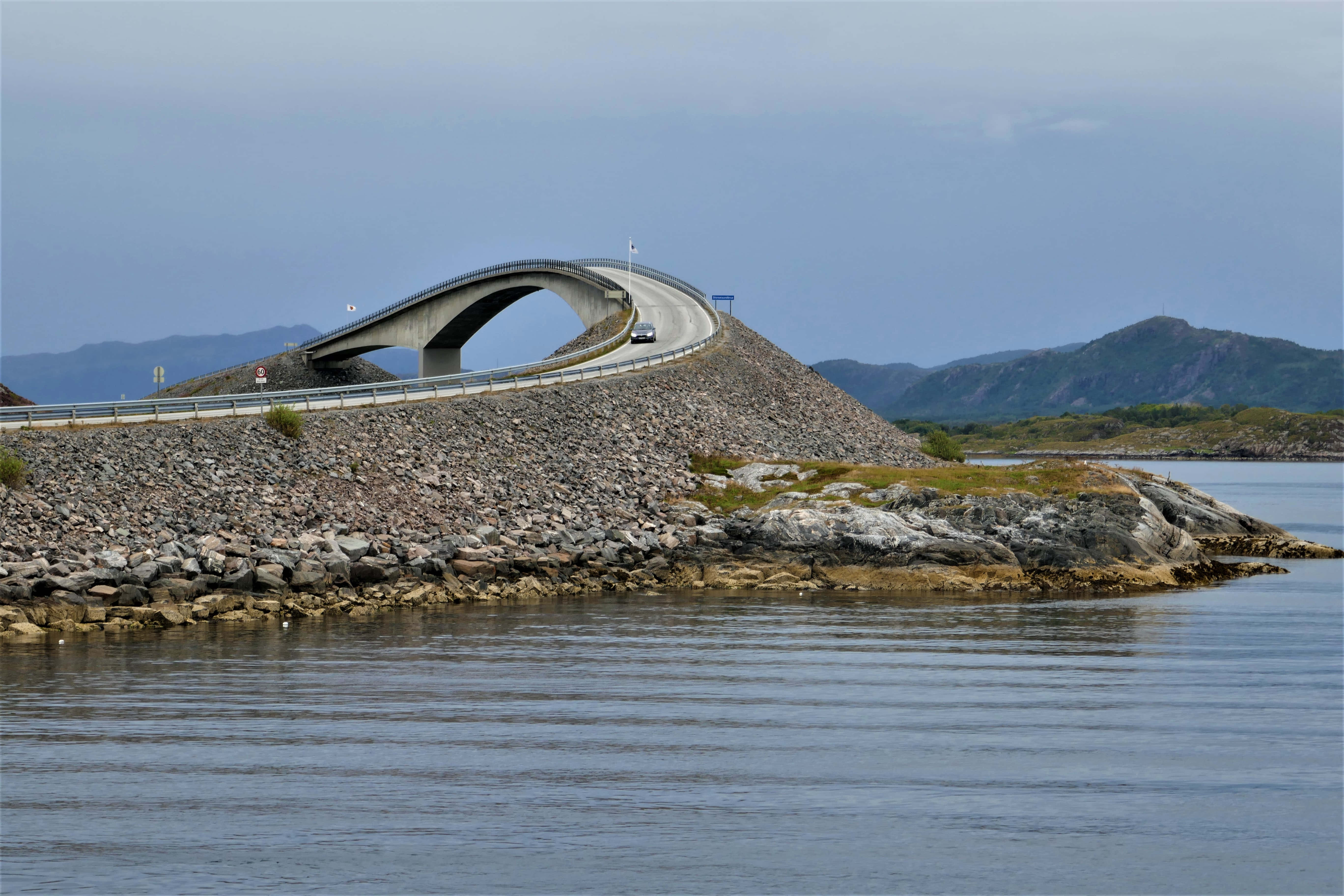 Road 桌布 壁紙 — 一座時尚的現代橋樑優雅地彎曲在平靜的水面上，遠山在陰天之下。