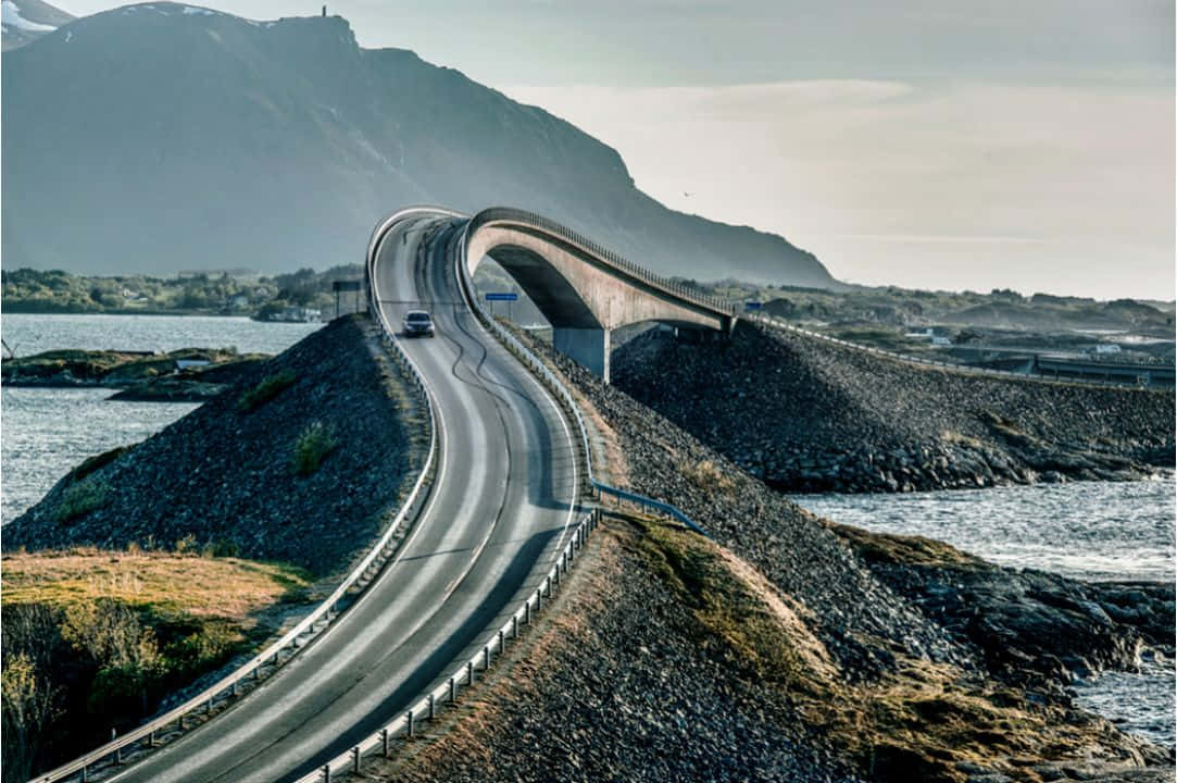 Road 桌布 壁紙 — 一座時尚的現代橋樑優雅地彎曲在水面上，一輛孤獨的汽車沿著蜿蜒的道路行駛，兩旁是岩石海岸和遠處的山脈。