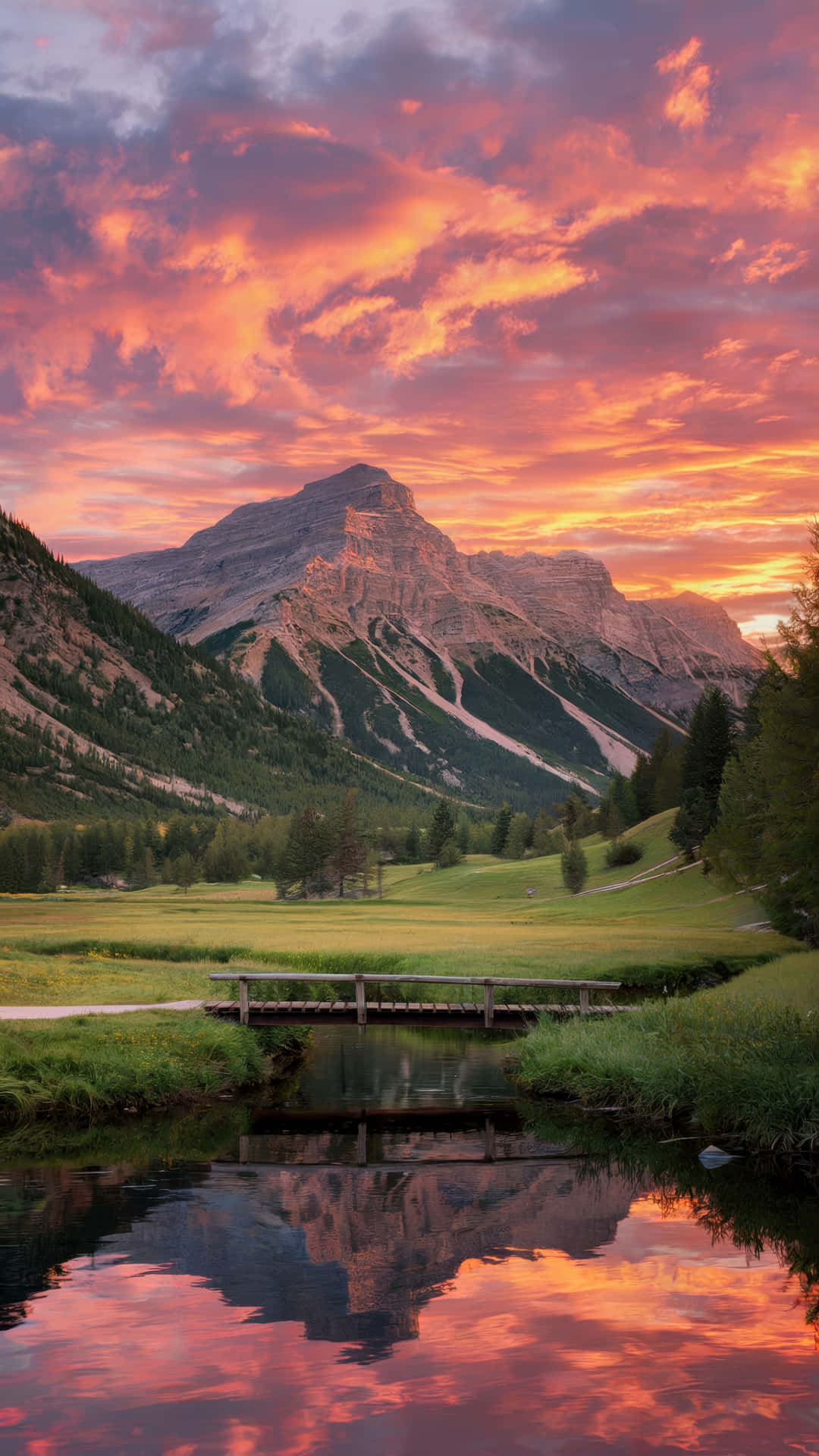 Majestic mountain landscape with vibrant sunset sky reflection 壁紙 — 一座木橋橫跨平靜的小溪，倒映著高聳的山峰，在火紅橙色和粉紅色的日落天空下。