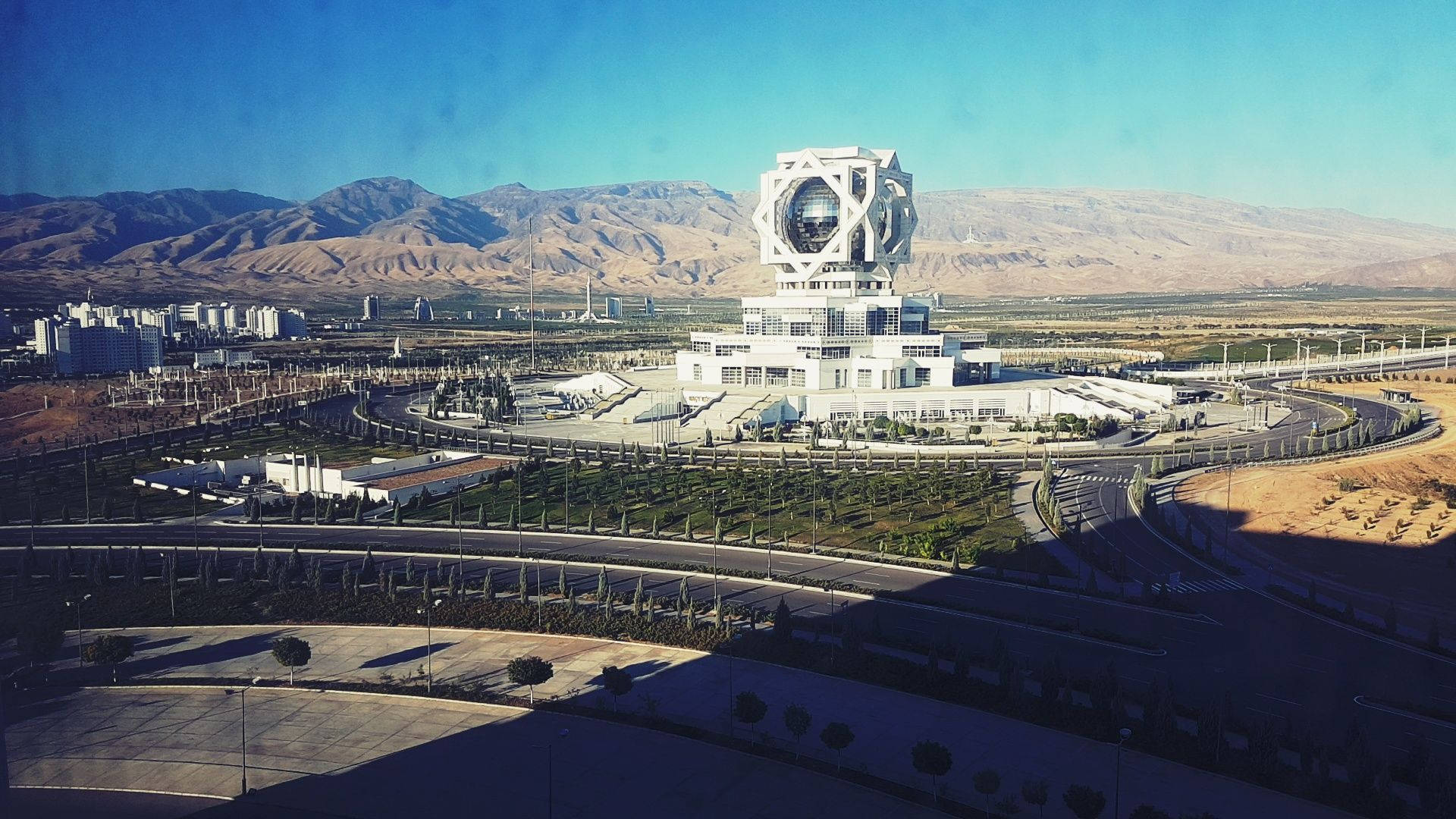Turkmenistan 桌布 壁紙 — 一座未來主義的白色建築，帶有球形中心，矗立在晴朗藍天下的沙漠景觀中。