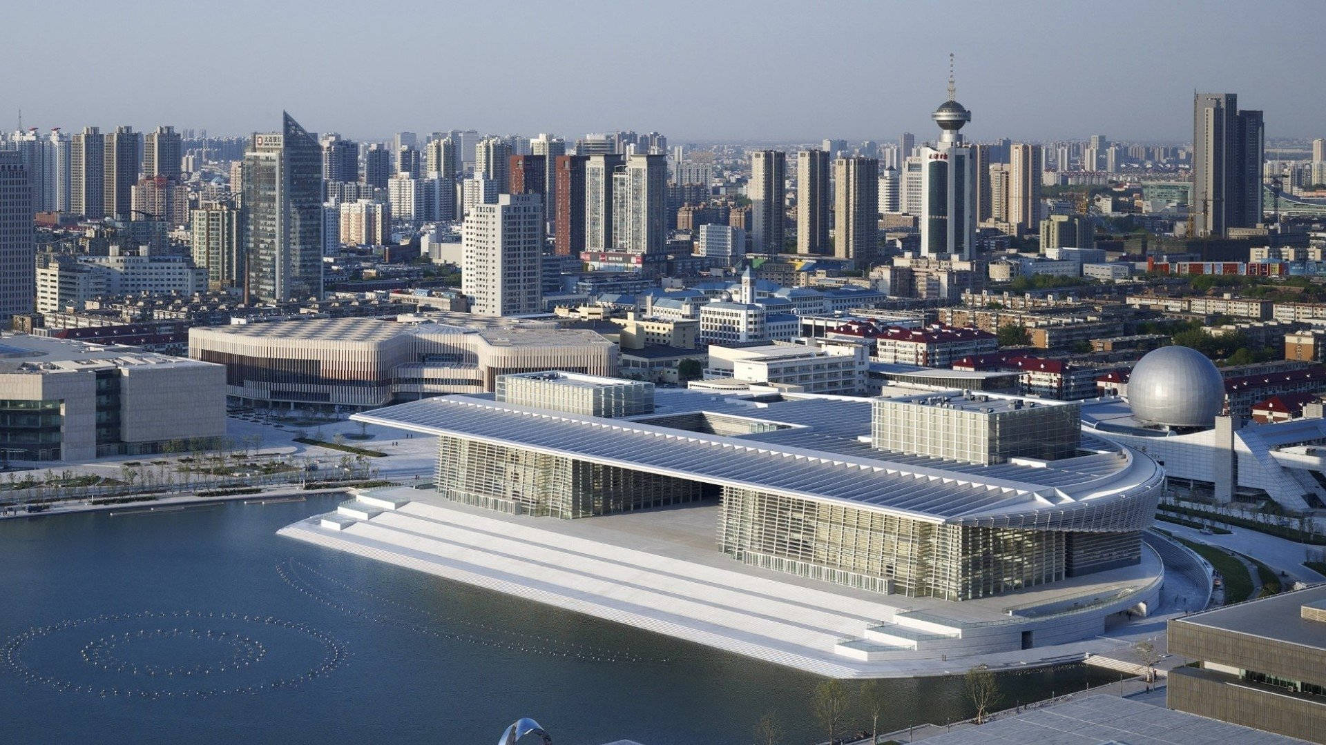 Tianjin 桌布 壁紙 — 一座未來主義風格的白色建築群，採用玻璃幕牆，坐落在晴朗天空下廣闊的現代城市景觀中，旁邊是平靜的水景。
