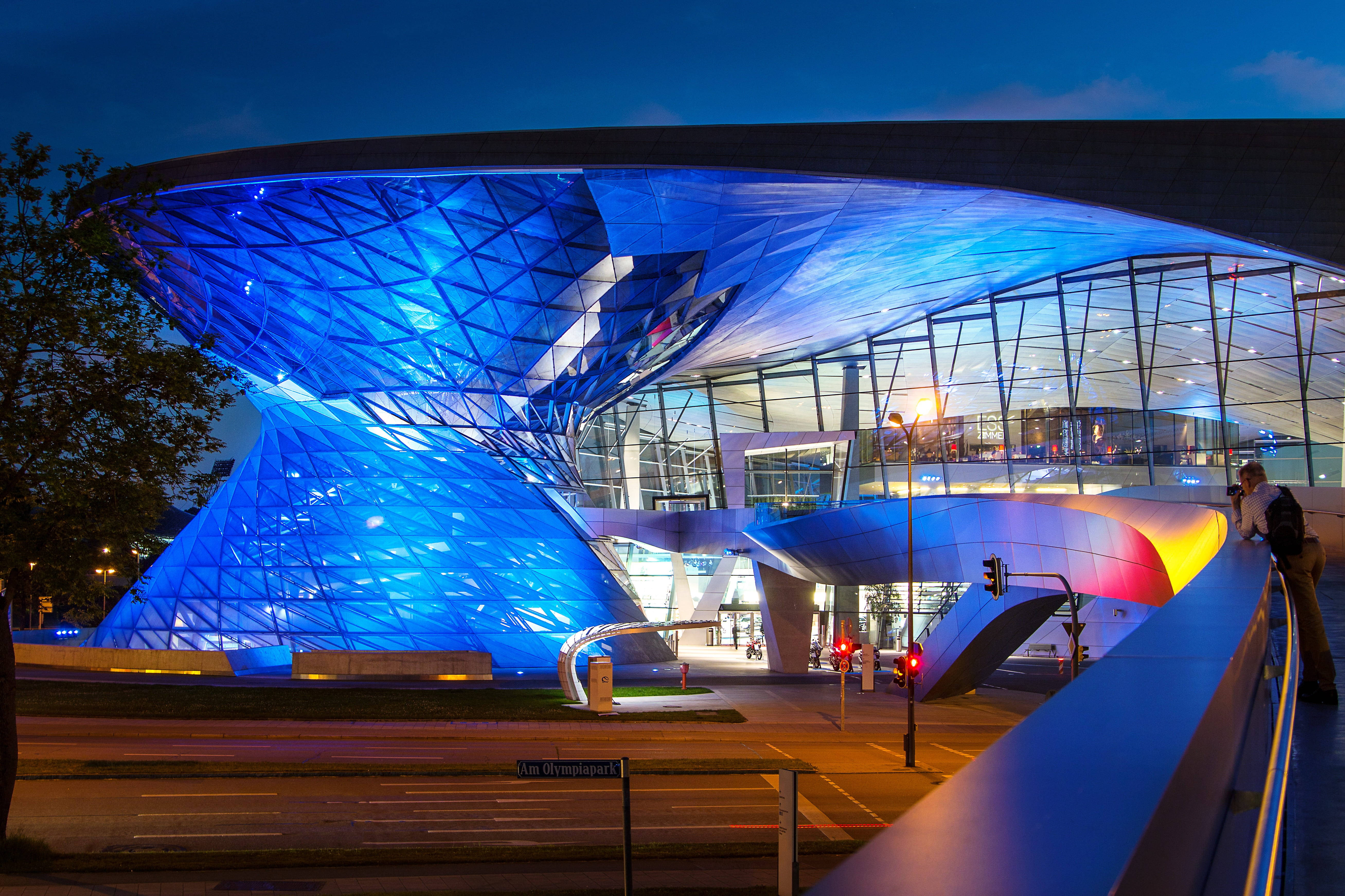 Munich 桌布 壁紙 — 一座未來感的藍色發光BMW Welt建築在夜晚閃耀，展現動態曲線和玻璃建築風格。