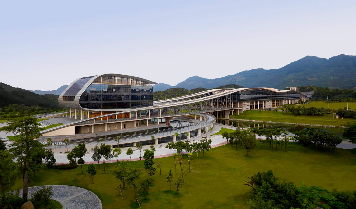 Shenzhen 桌布 壁紙 — 一座未來風格的玻璃和鋼結構建築，採用弧形設計，坐落在茂密的綠色草坪和遠處山脈之間，天空晴朗。