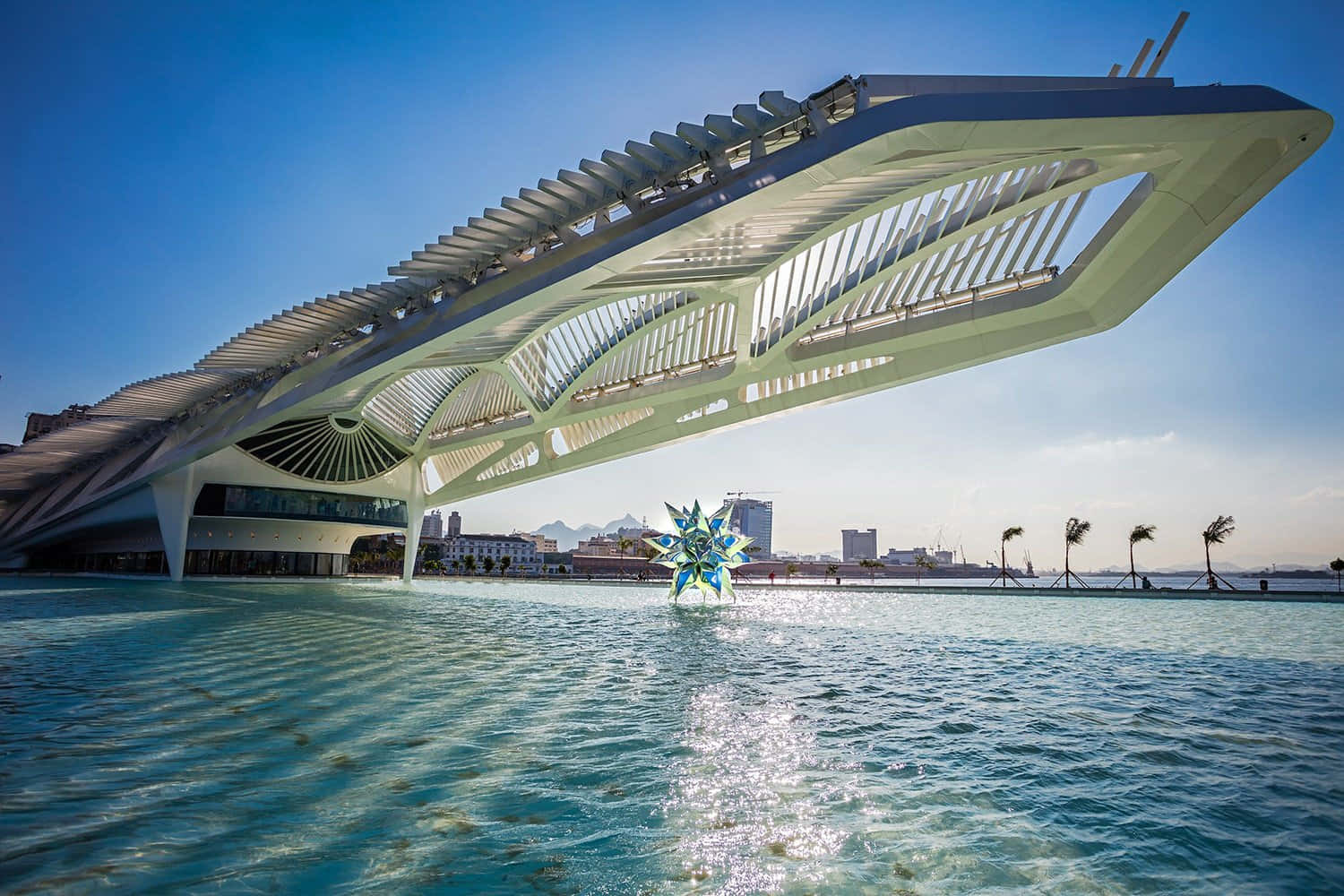 Beautiful Architecture 桌布 壁紙 — 一座未來風格的白色建築橋樑拱跨在碧綠色的水面上，背景是城市天際線和棕櫚樹，在清澈的藍天下。