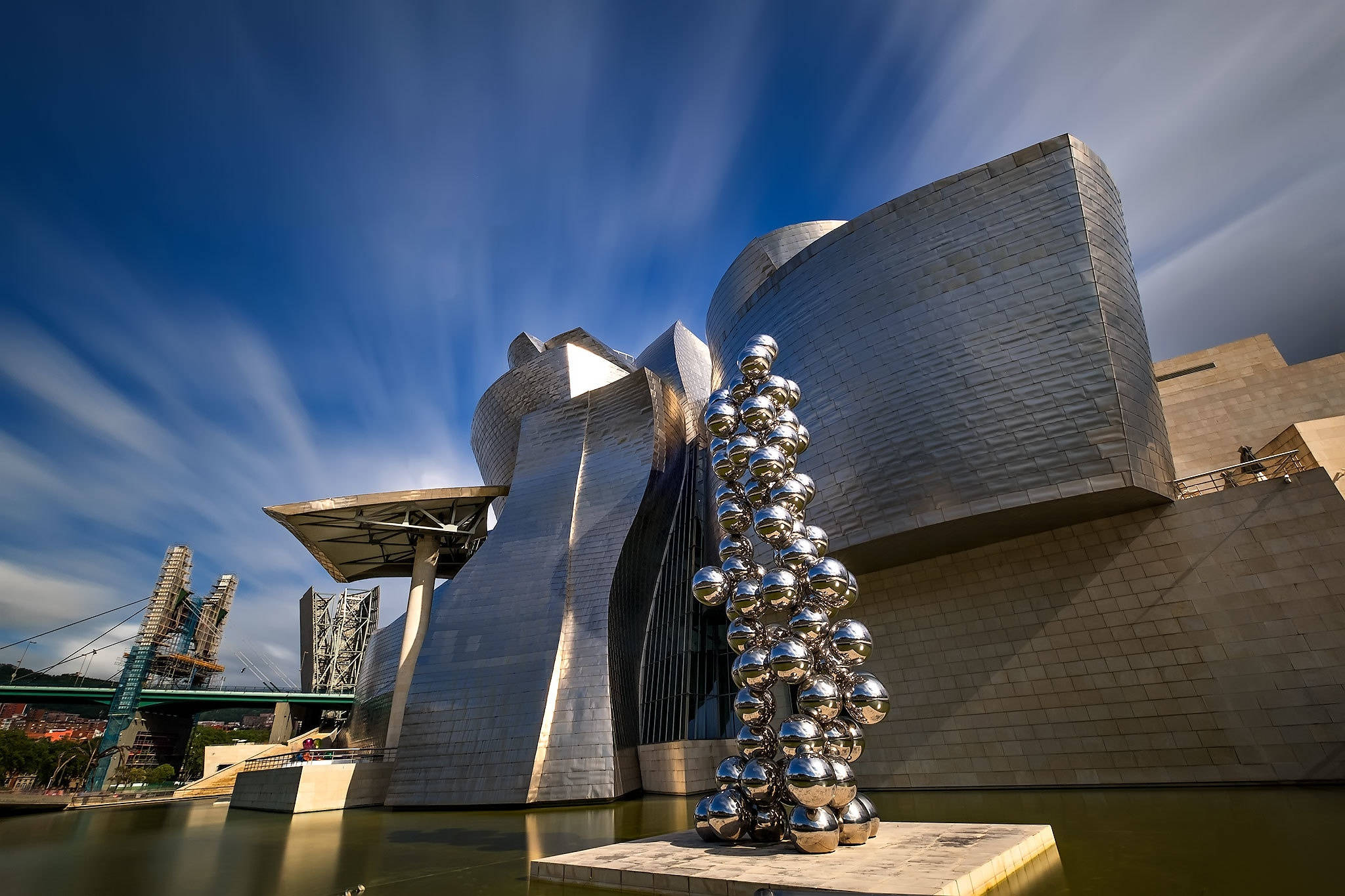 Museum 桌布 壁紙 — 一座未來風格的銀色建築，帶有彎曲的金屬牆壁和一個反射球體雕塑，矗立在水邊，上方是帶有條紋雲朵的鮮豔藍天。