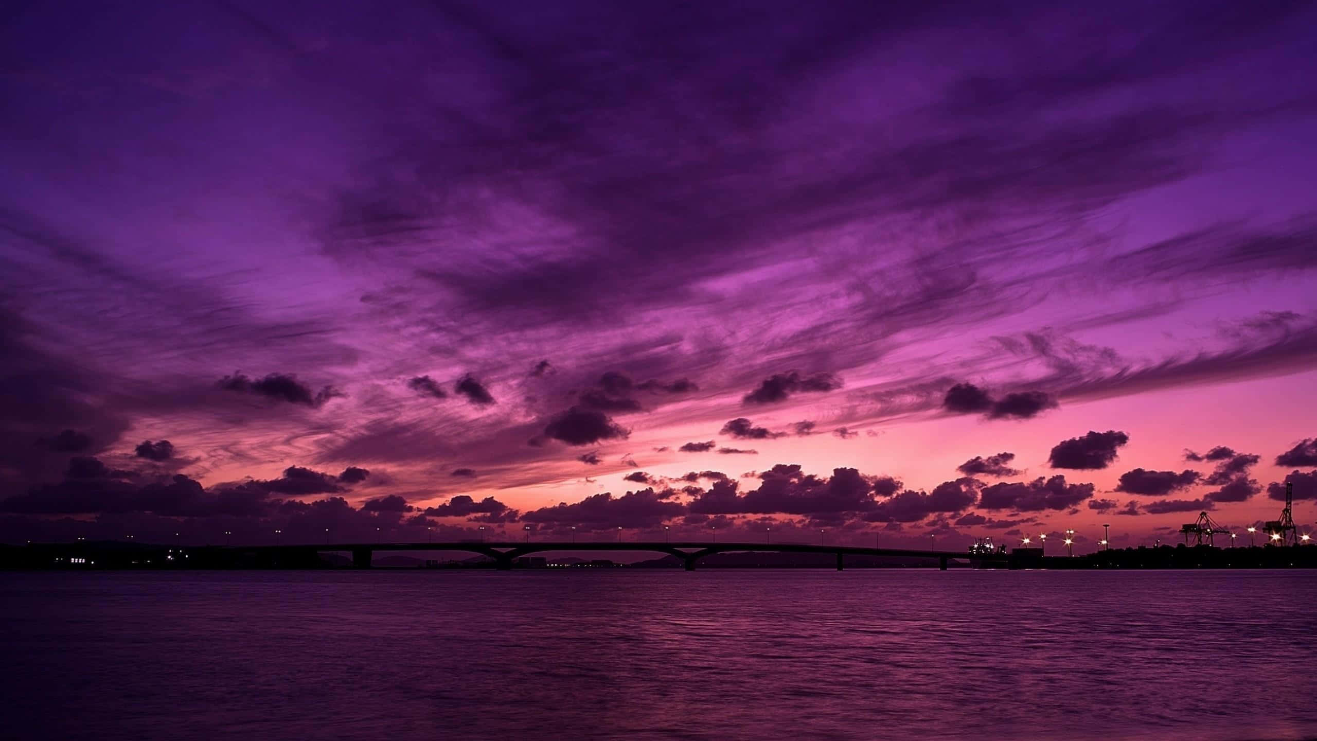 Purple Aesthetic Phone 桌布 壁紙 — 一座橋樑橫跨平靜的水道，天空呈現戲劇性的紫色和粉紅色日落，雲朵散佈。