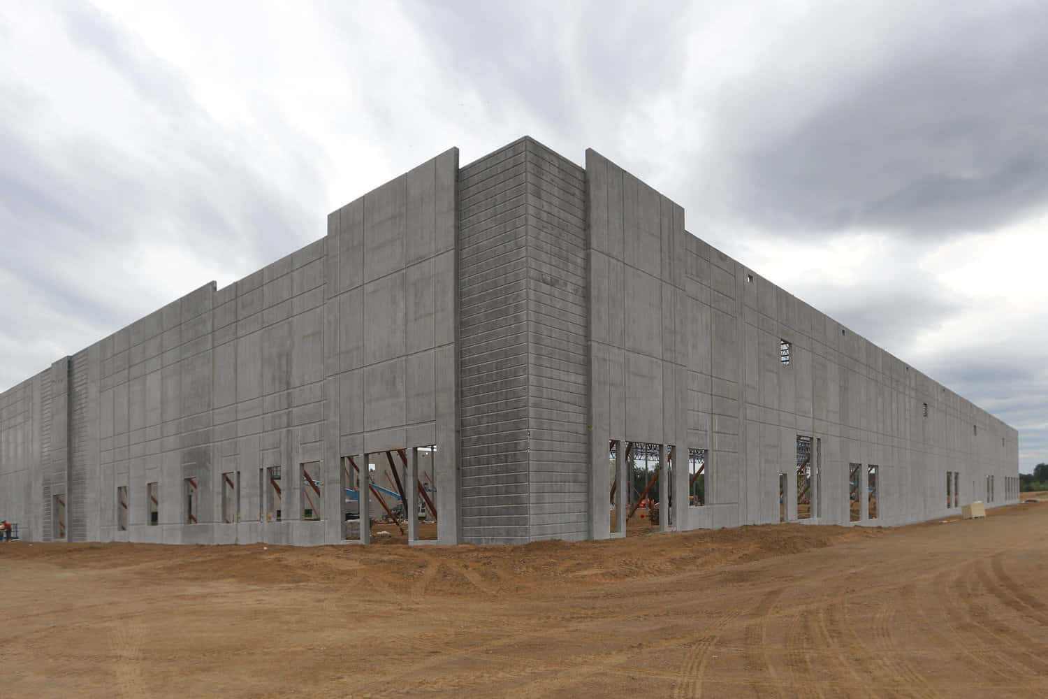 建築 桌布 壁紙 — 一座正在建造中的巨大未完成混凝土建築，帶有暴露的結構框架和泥濘的地面，天空多雲。