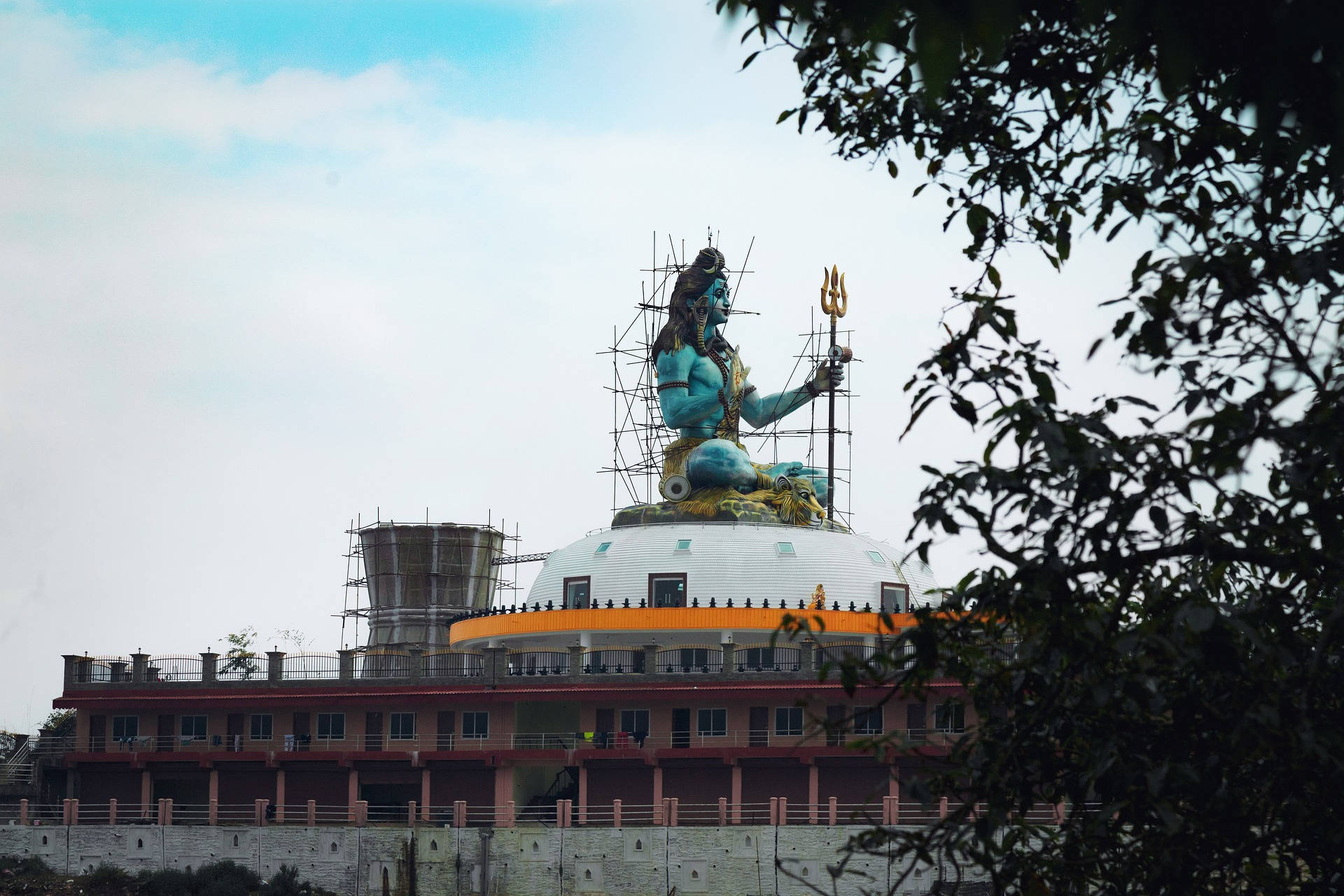 shiv shankar 桌布 壁紙 — 一座正在建造中的高聳藍色濕婆神像，矗立於圓頂寺廟之上，以多雲的天空為背景，被茂密的樹枝環繞。