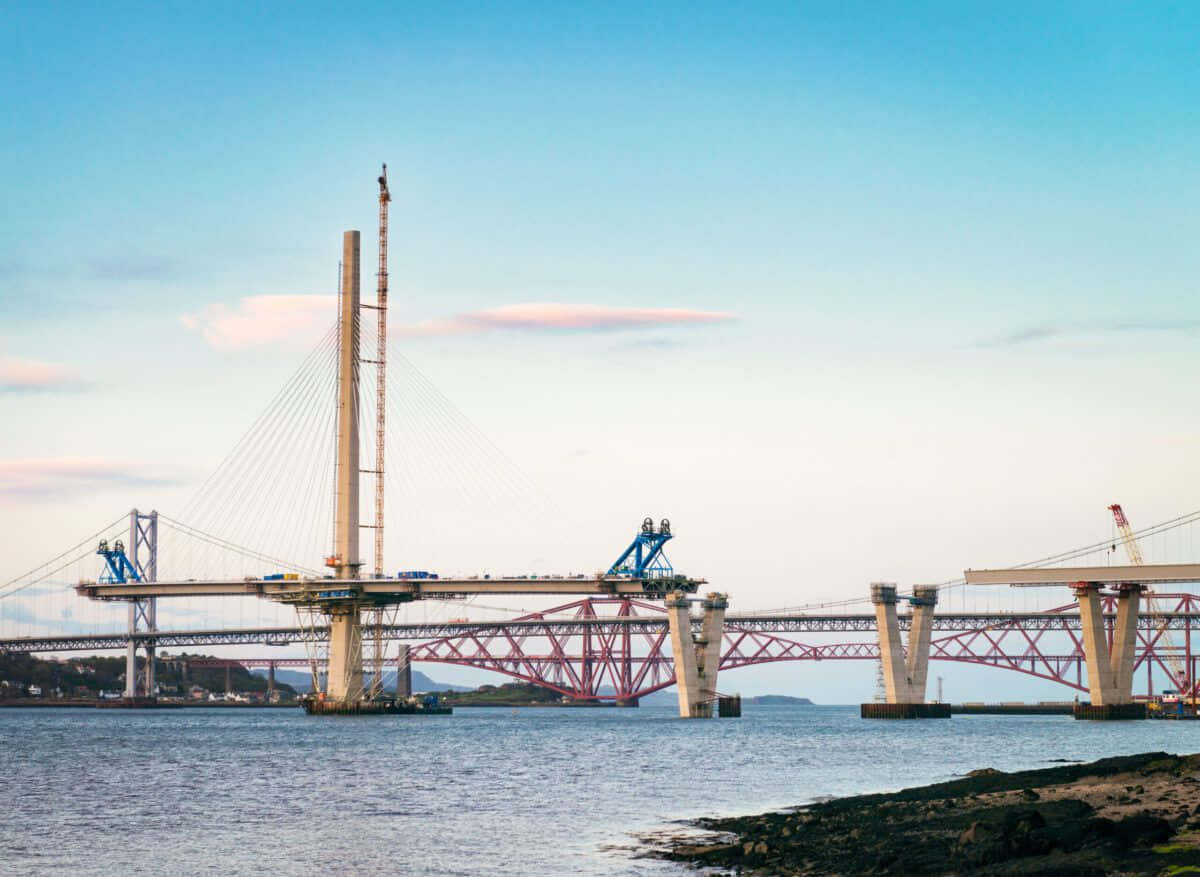 Civil Engineering 桌布 壁紙 — 一座正在建造的現代斜張橋橫跨寬闊的河流，背景中一座歷史悠久的紅色懸臂橋在晴朗的藍天下清晰可見。