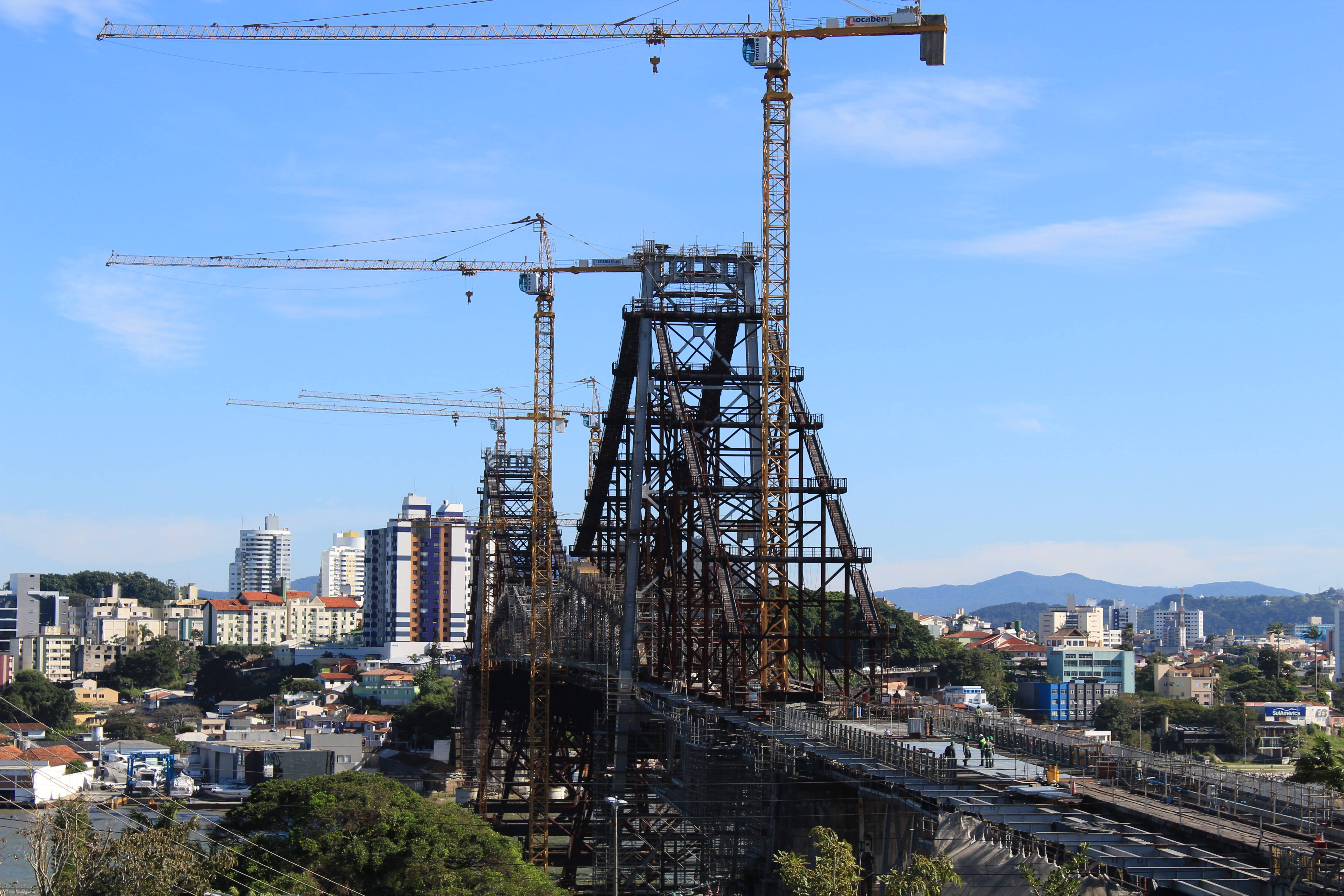 建築 桌布 壁紙 — 一座正在建造的高聳鋼橋，在晴朗的藍天下，主導著一個充滿起重機和現代建築的繁華城市景觀。