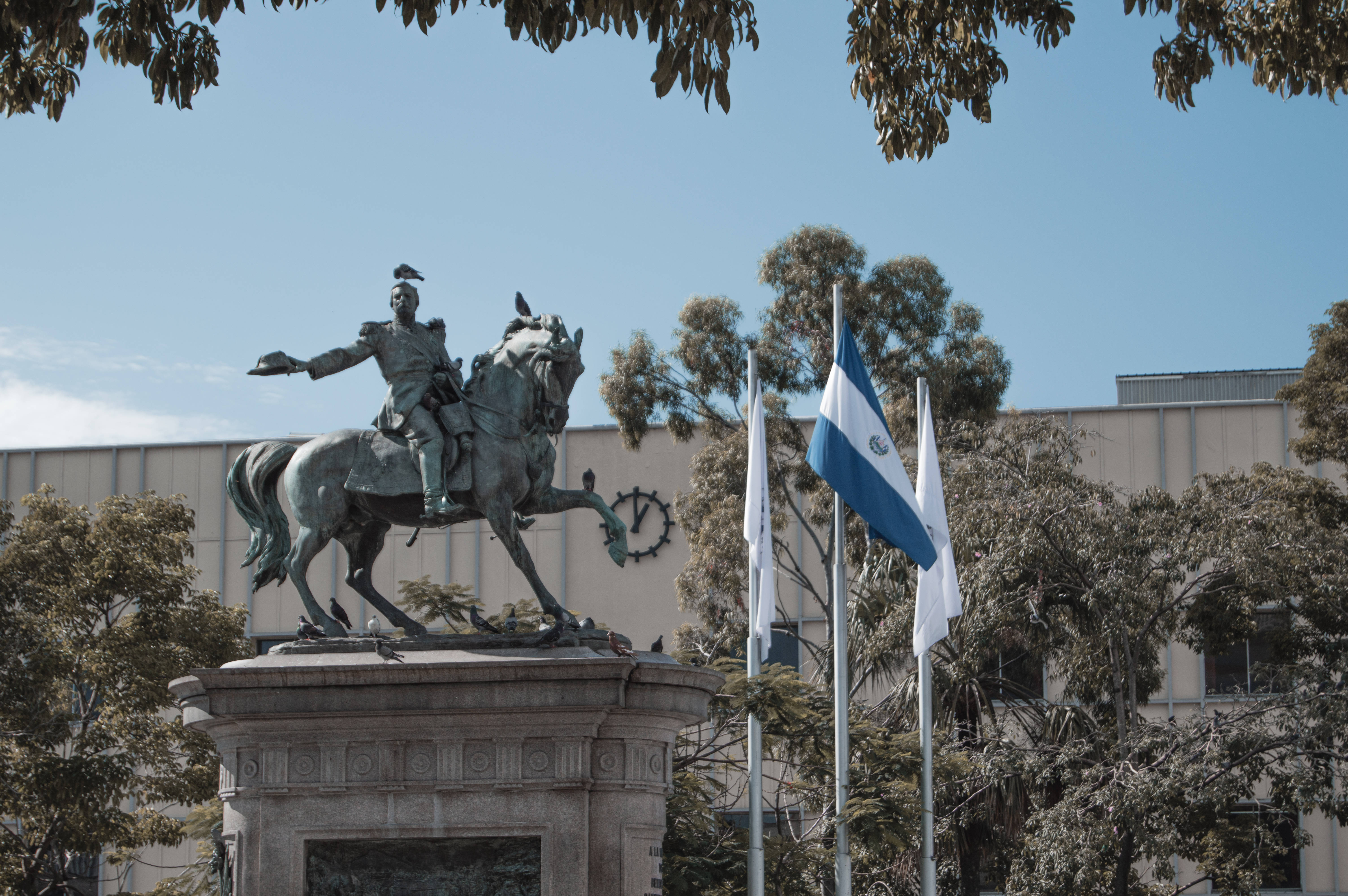 El Salvador 桌布 壁紙 — 一座歷史人物的青銅騎馬雕像自豪地矗立在石基座上，背景是晴朗的藍天，附近有樹木和國旗。