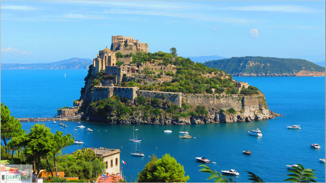Italian Island 桌布 壁紙 — 一座歷史悠久的堡壘坐落在一個鬱鬱蔥蔥的綠色島嶼上，周圍是湛藍的海水，點綴著船隻，天空晴朗。