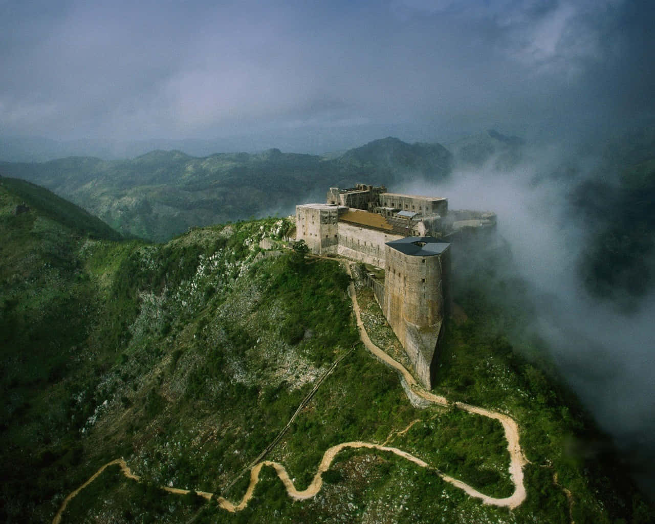 Fortress 桌布 壁紙 — 一座歷史悠久的堡壘坐落在霧氣繚繞的山脊上，蜿蜒的泥土小徑穿過下方鬱鬱蔥蔥的綠色山谷。