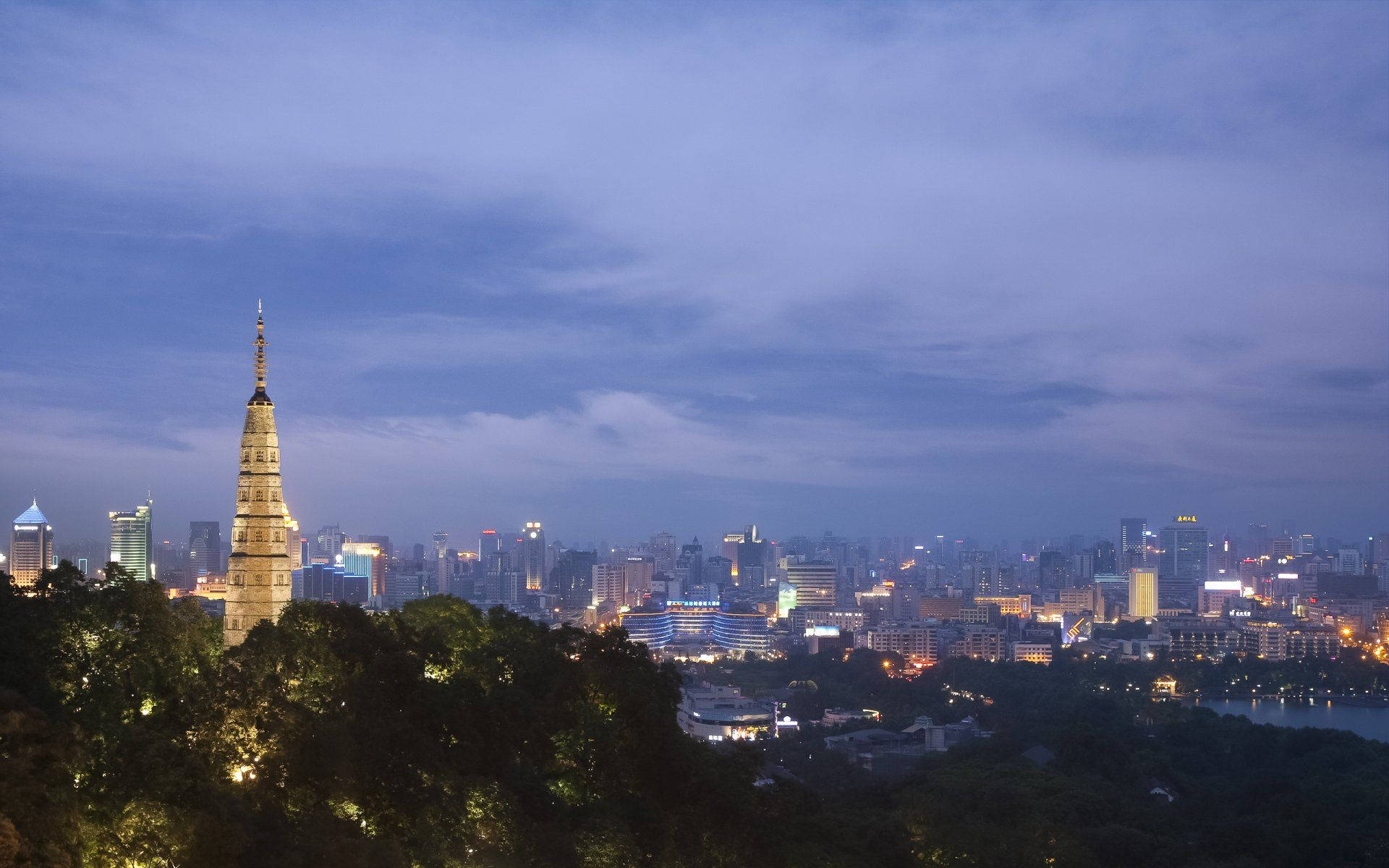 Hangzhou 桌布 壁紙 — 一座歷史悠久的寶塔在樹木繁茂的山頂上閃耀著光芒，俯瞰著暮色中廣闊的城市景觀，伴隨著柔和的紫色雲彩。