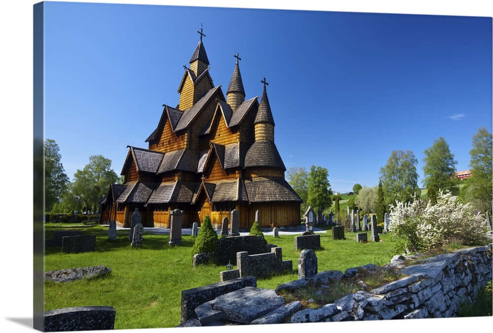 Norway 桌布 壁紙 — 一座歷史悠久的木造木板教堂，有多個尖頂，矗立在晴朗的藍天下，周圍是綠色的墓地，有石頭墳墓和盛開的白花。