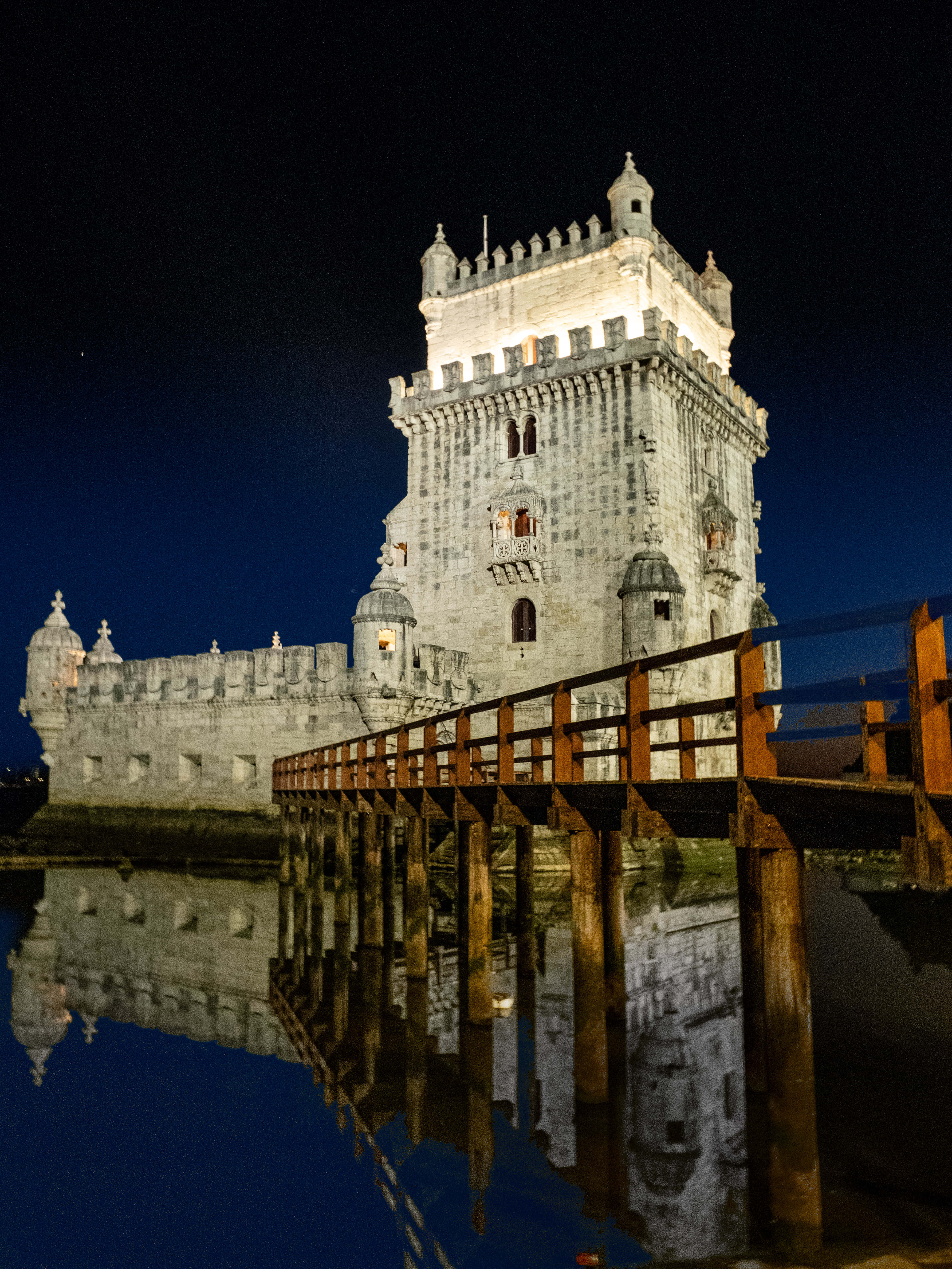 Portugal 桌布 壁紙 — 一座歷史悠久的石塔在夜晚被照亮，其倒影完美地映照在木碼頭下的平靜水面上。